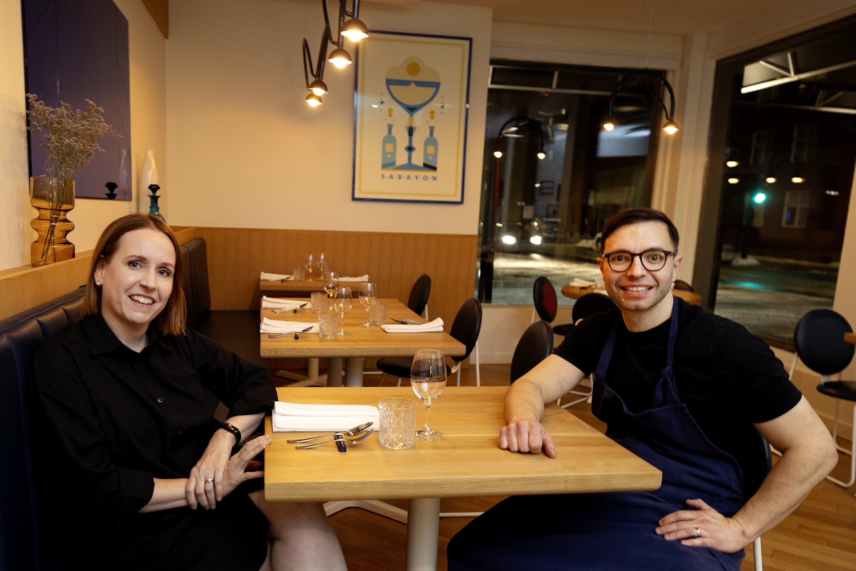 A woman and man on opposite sides of a table in an empty restaurant smile at the camera.