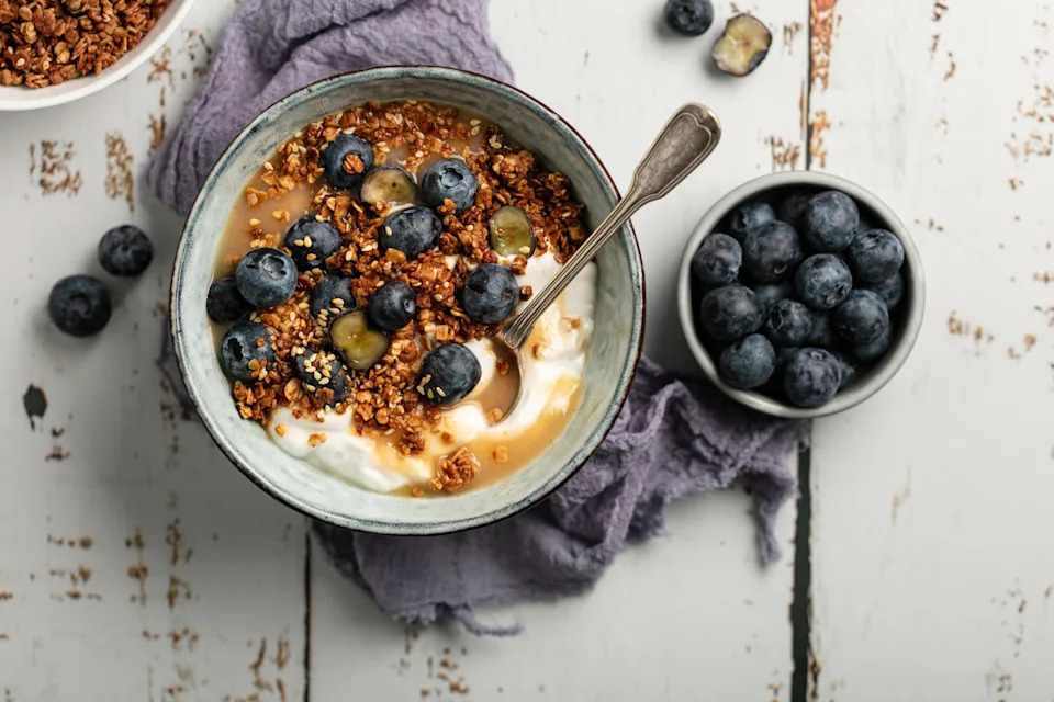 A bowl of yogurt topped with granola and blueberries on a rustic table, with additional blueberries in a small dish beside it