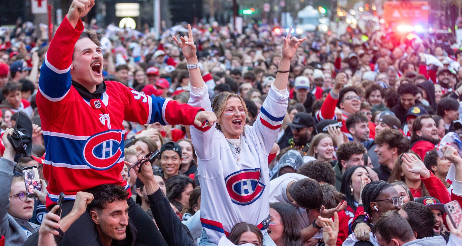 A crowd of cheering hockey fans. Two people in Habs jerseys, a man and woman, are on people's shoulders and waving their hands in the air.