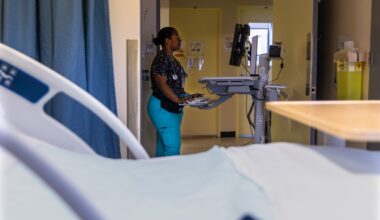 Nurse typing into a a computer on a portable workstation with hospital bed in the foreground