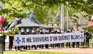 A line of masked men stand in a park holding a sign that reads Je me souviens d'un Québec blanc.