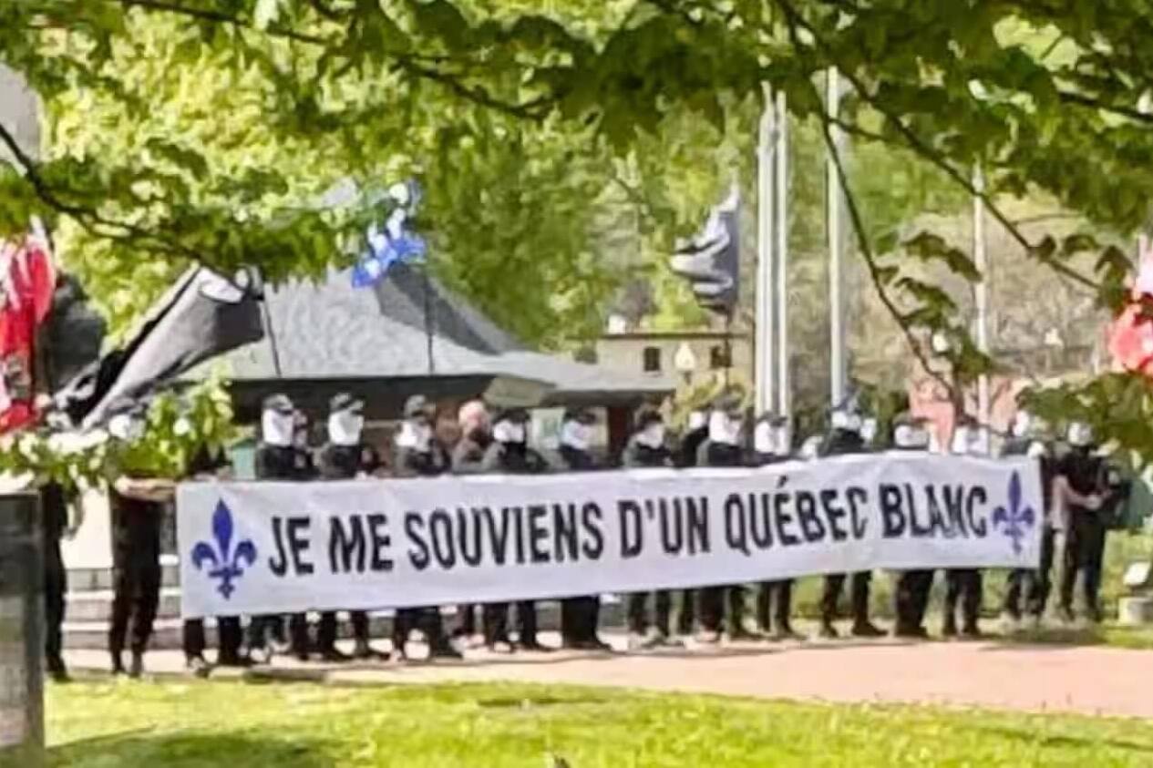 A line of masked men stand in a park holding a sign that reads Je me souviens d'un Québec blanc.