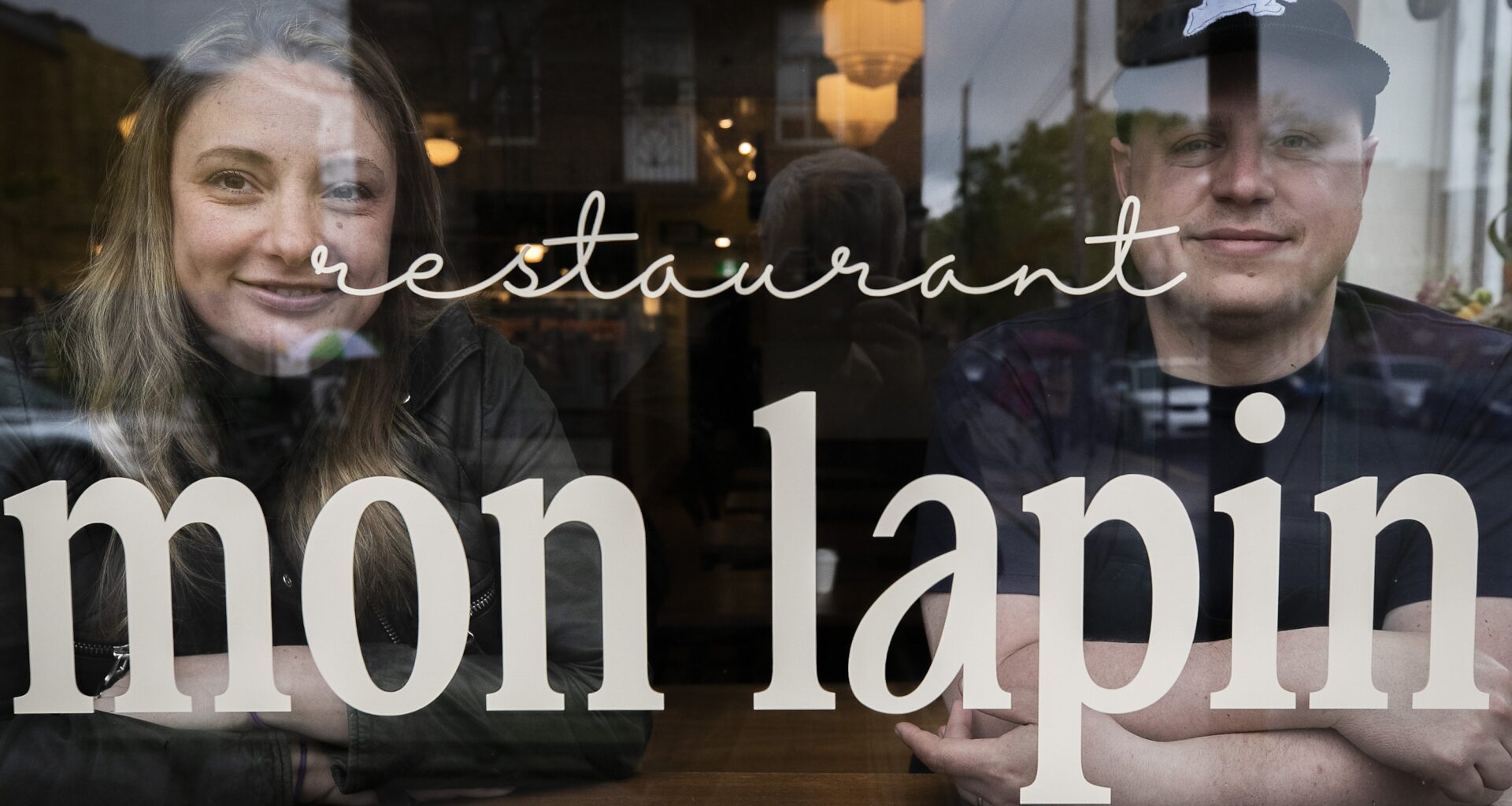 A woman and man sit behind a restaurant window that has the words Restaurant Mon Lapin on it.