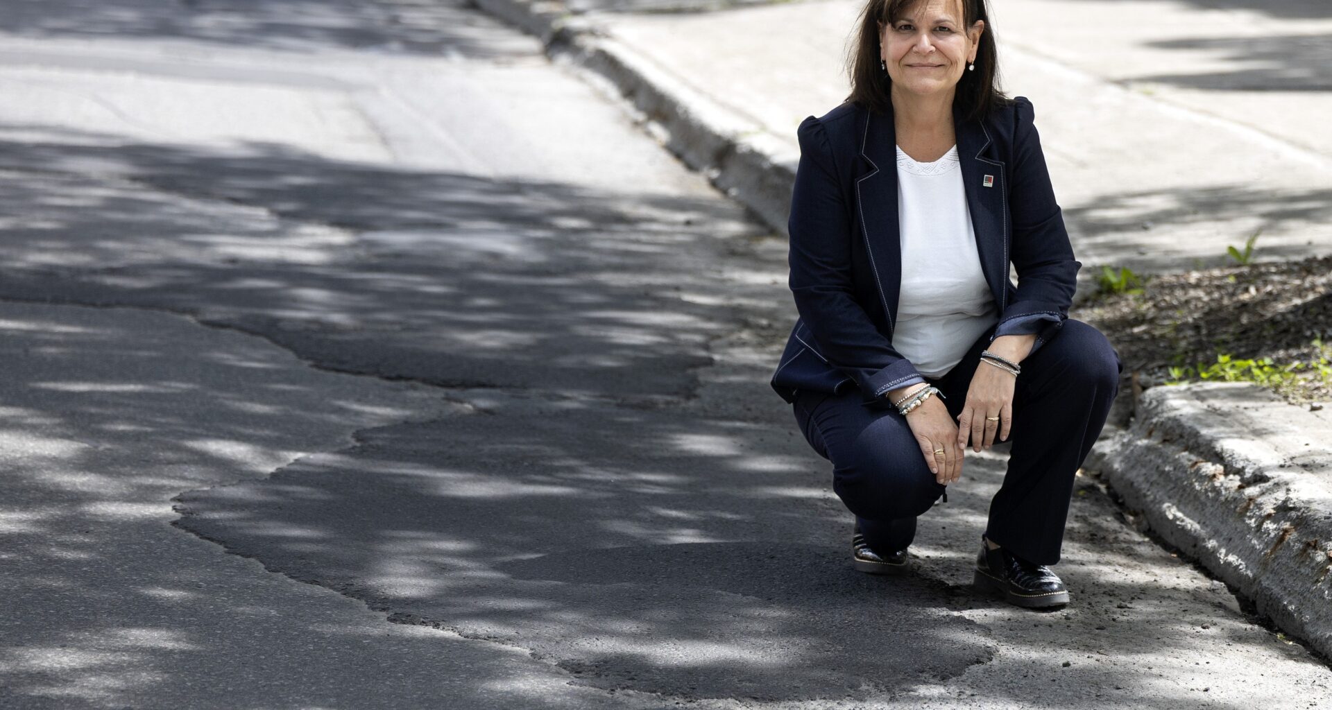 A woman crouches down at the side of a road marked with pothole repairs.