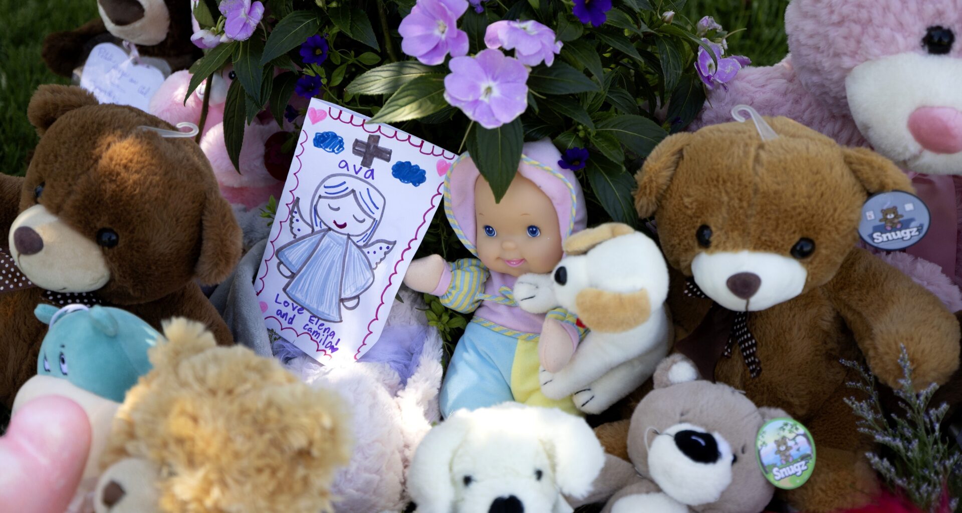 A memorial made up of plush animals, toys and cards is seen among greenery. A card depicts a girl as an angel, with the name Ava written on it.