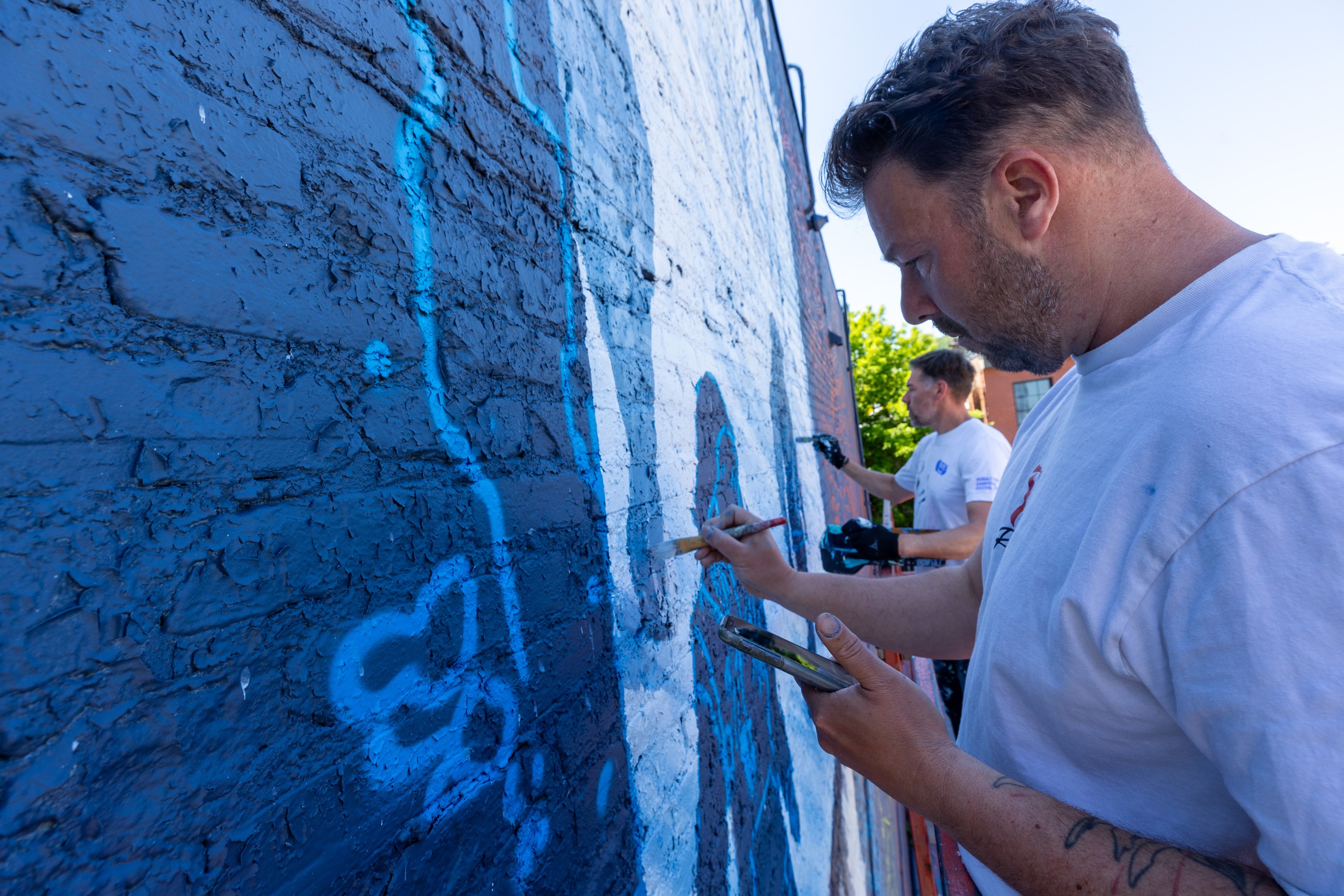 Two men work on a large mural on the side of a building, painting in blue.
