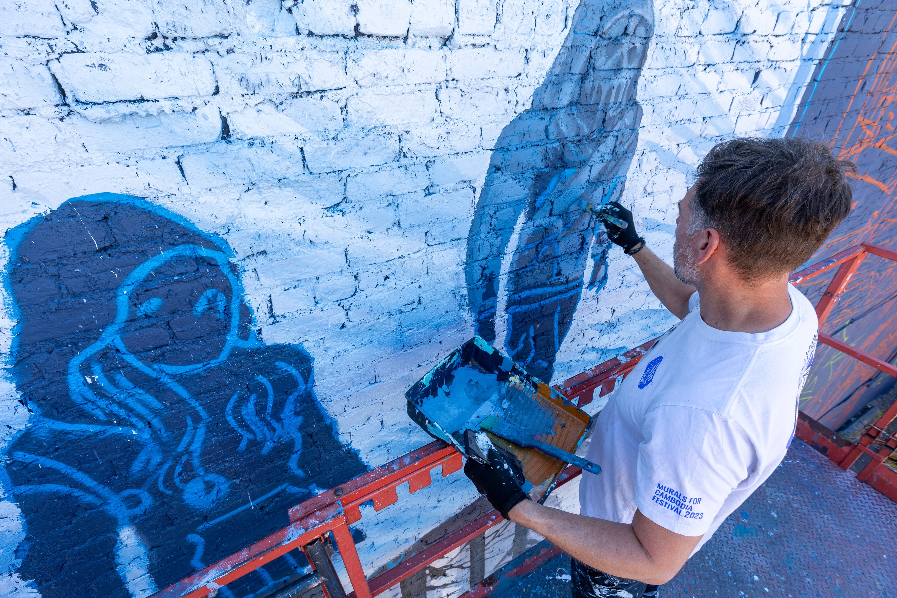 A man is seen from above while working on a mural on the side of a building, painting a figure in blue.