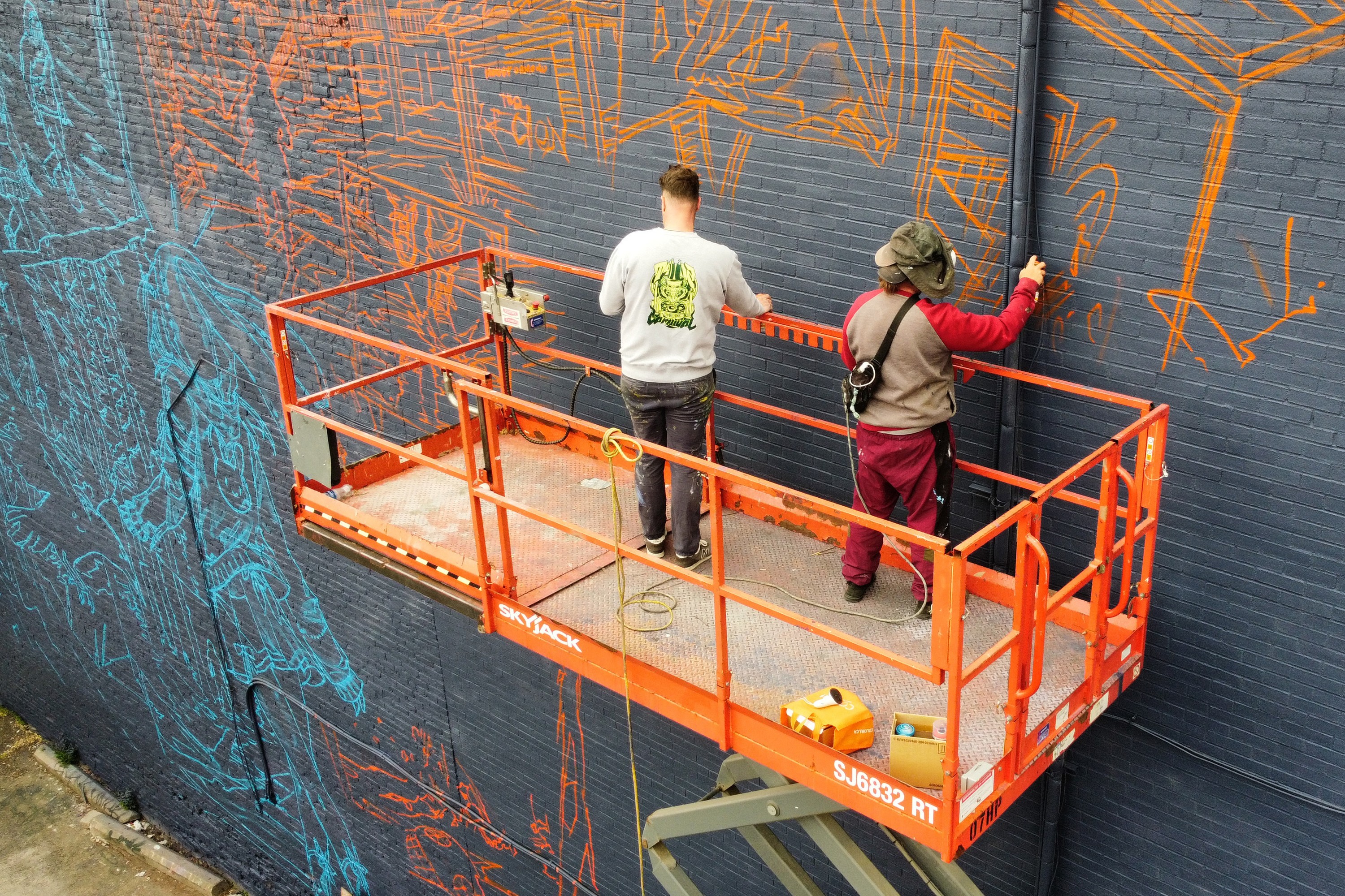 Two people on a cherry picker sketch orange and blue drawings on a tall wall. One is wearing a VR headset.