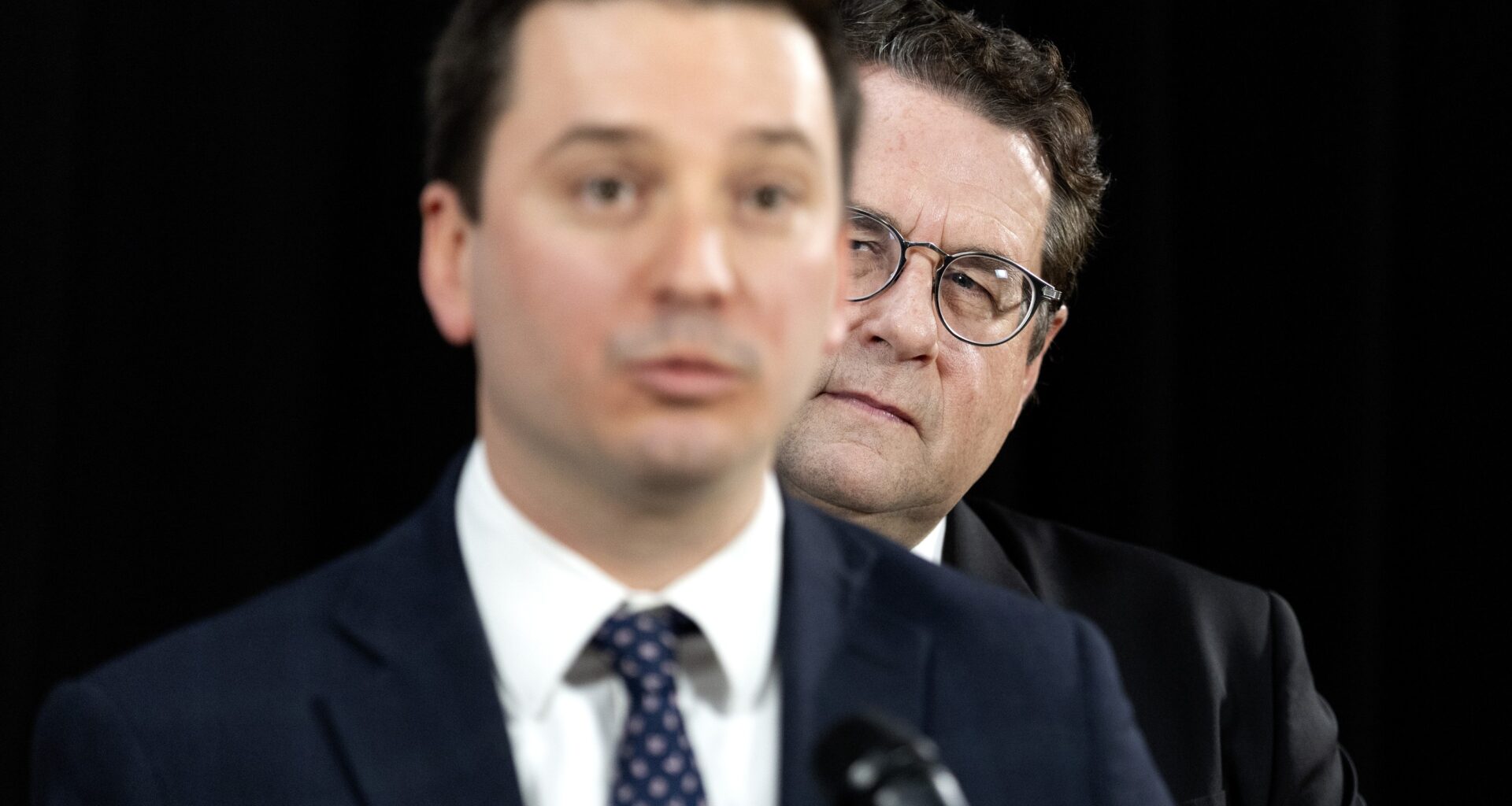 Simon Jolin-Barrette, out of focus, speaks at a mic as Bernard Drainville, in focus, looks from behind