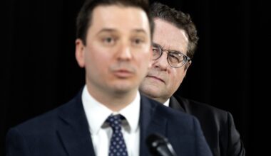 Simon Jolin-Barrette, out of focus, speaks at a mic as Bernard Drainville, in focus, looks from behind