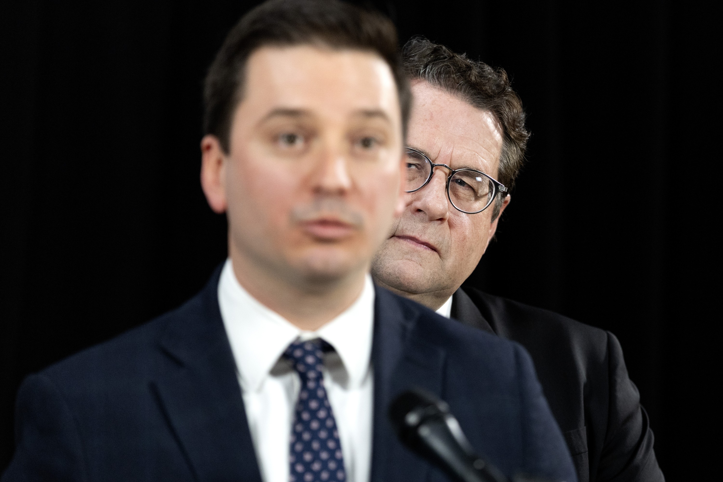 Simon Jolin-Barrette, out of focus, speaks at a mic as Bernard Drainville, in focus, looks from behind