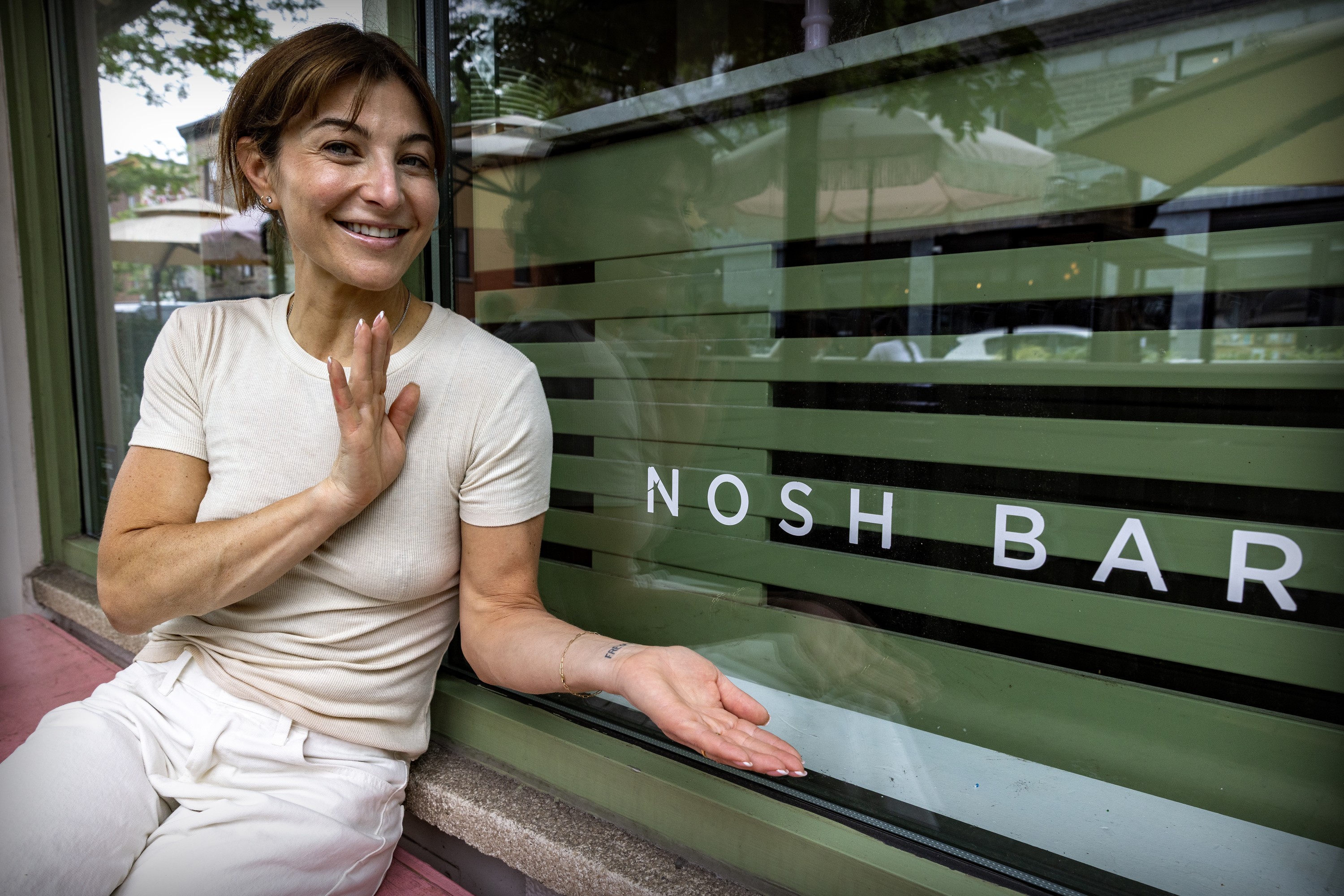 A smiling woman holds out her hands to show a sign on a window that says Nosh Bar