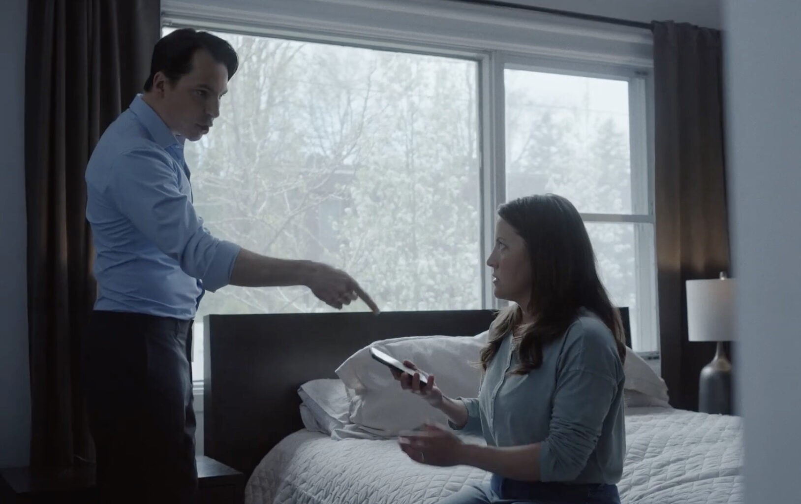 A man points menacingly at his partner as she sits on a bed, holding a phone.