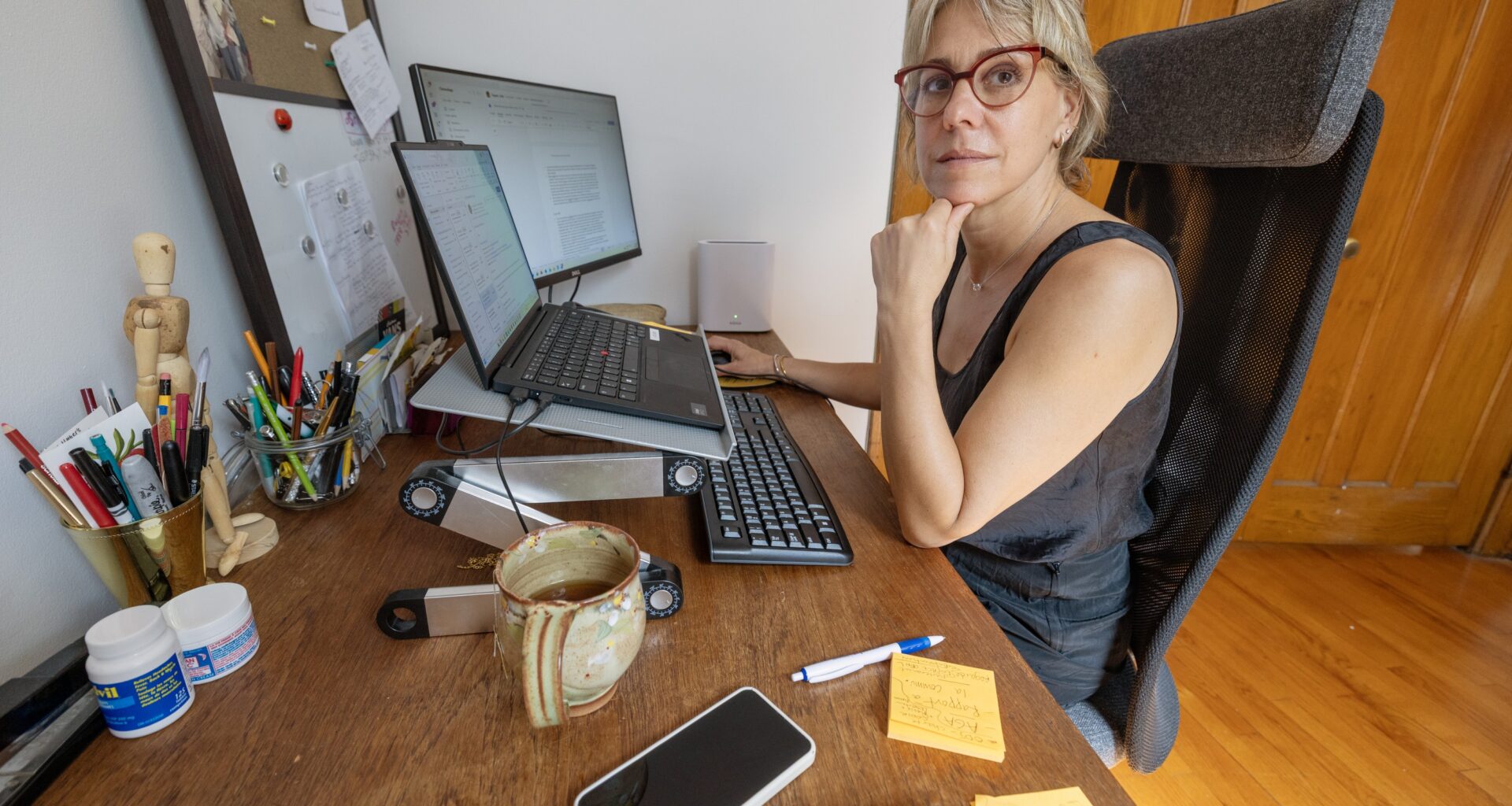 A woman sits at a desk in her home office. There are two monitors, a corkboard and cups of pens on the desk.