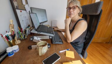 A woman sits at a desk in her home office. There are two monitors, a corkboard and cups of pens on the desk.