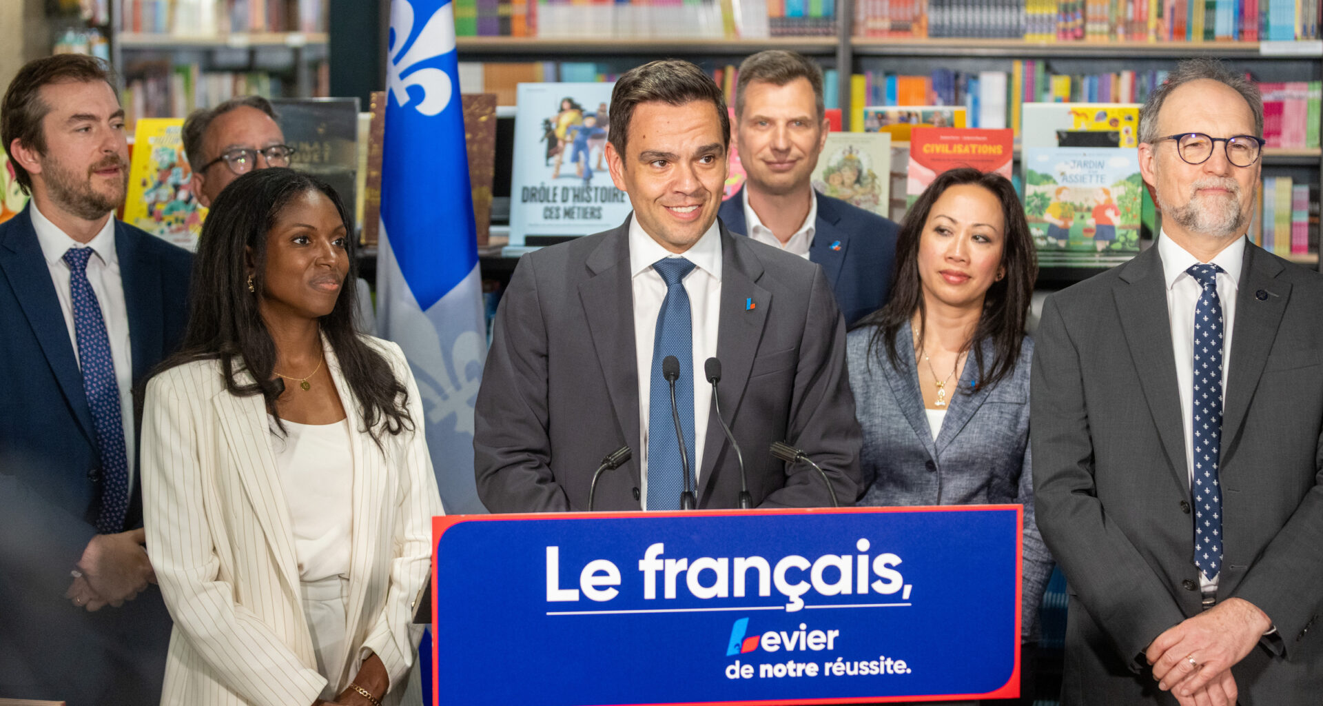 Charles Milliard and other people gather around a podium with shelves of books in the background.