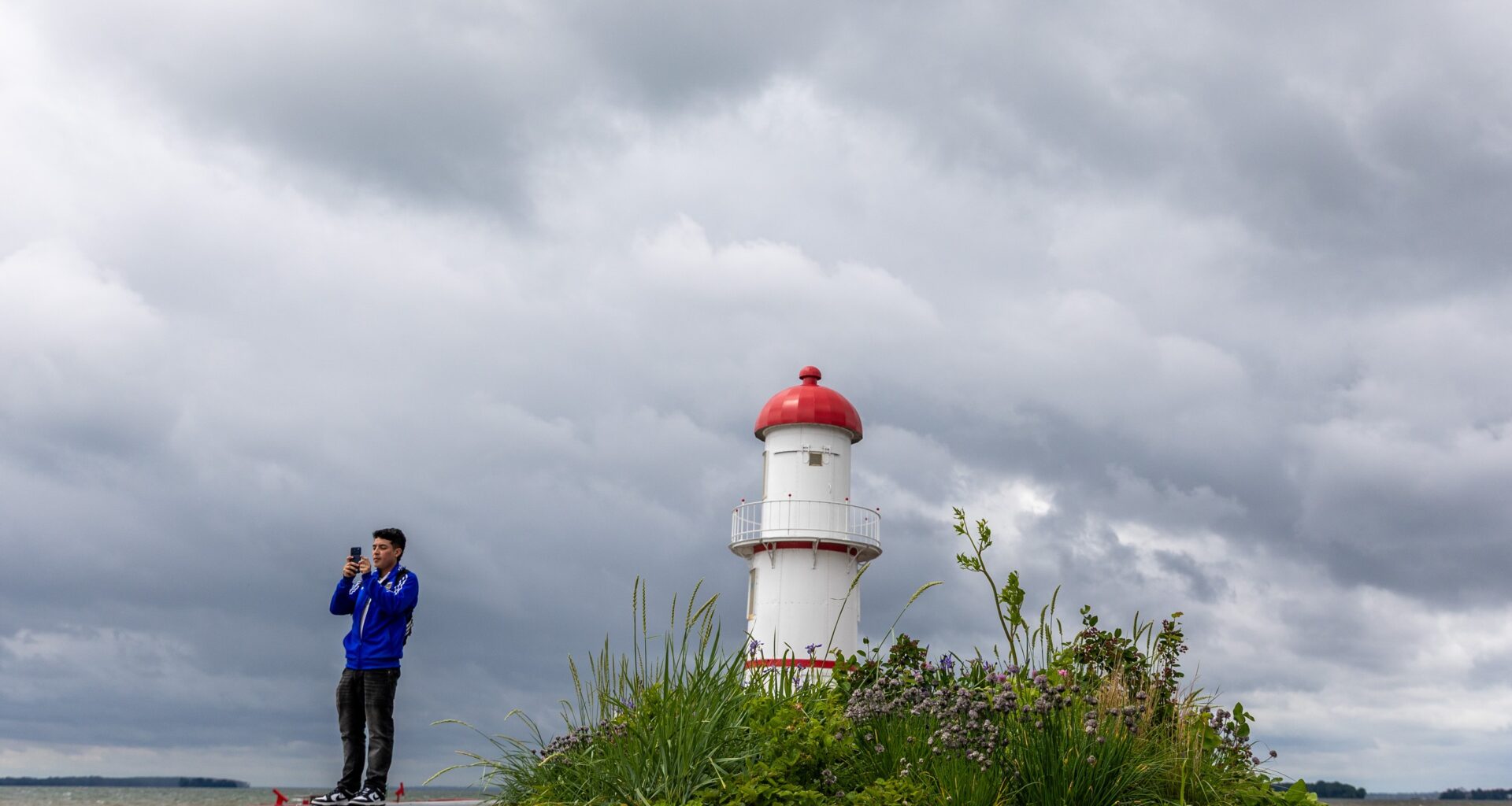 Under dark clouds, a person takes a photo of the shoreline with a lighthouse in the background.