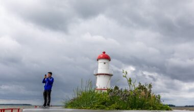 Under dark clouds, a person takes a photo of the shoreline with a lighthouse in the background.
