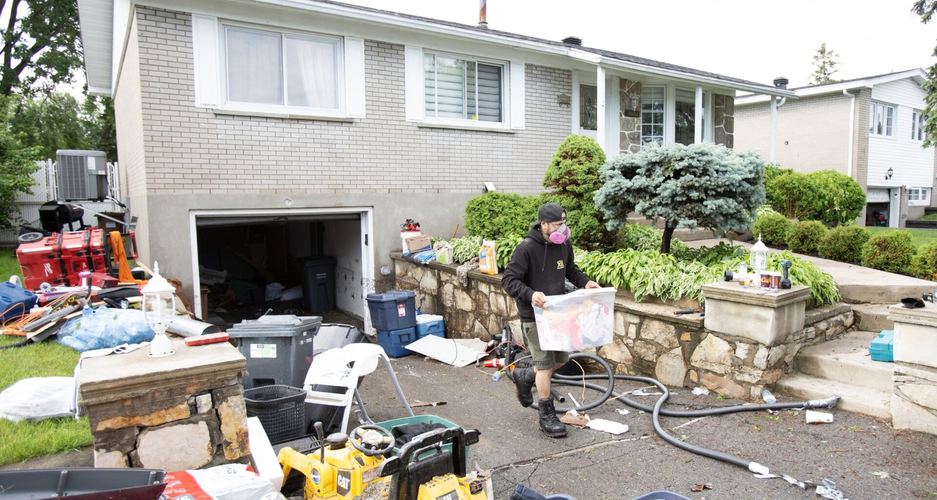 A man wearing a mask carries a container of belongings up a driveway strewn with belongings after flooding.
