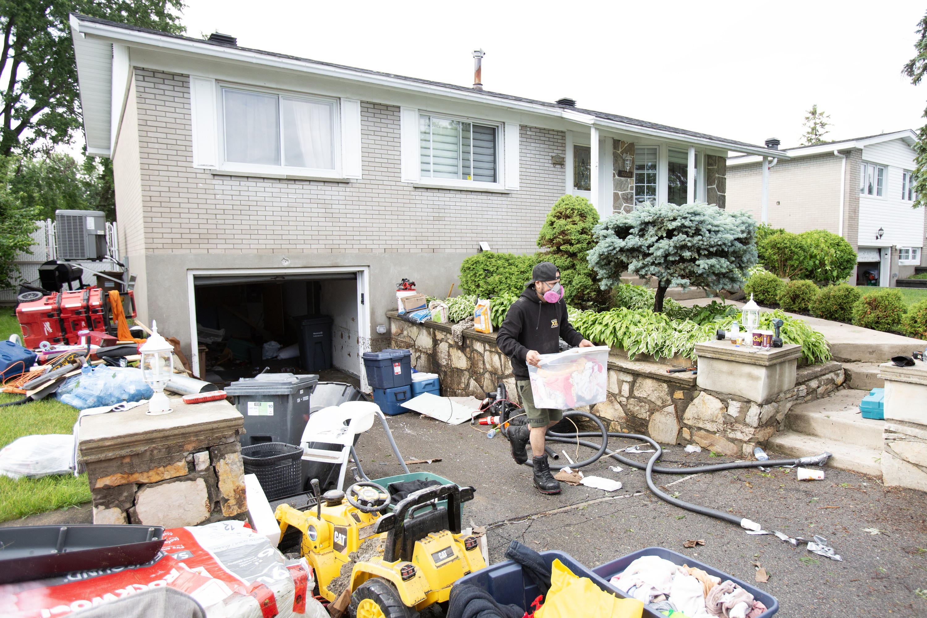 A man wearing a mask carries a container of belongings up a driveway strewn with belongings after flooding.