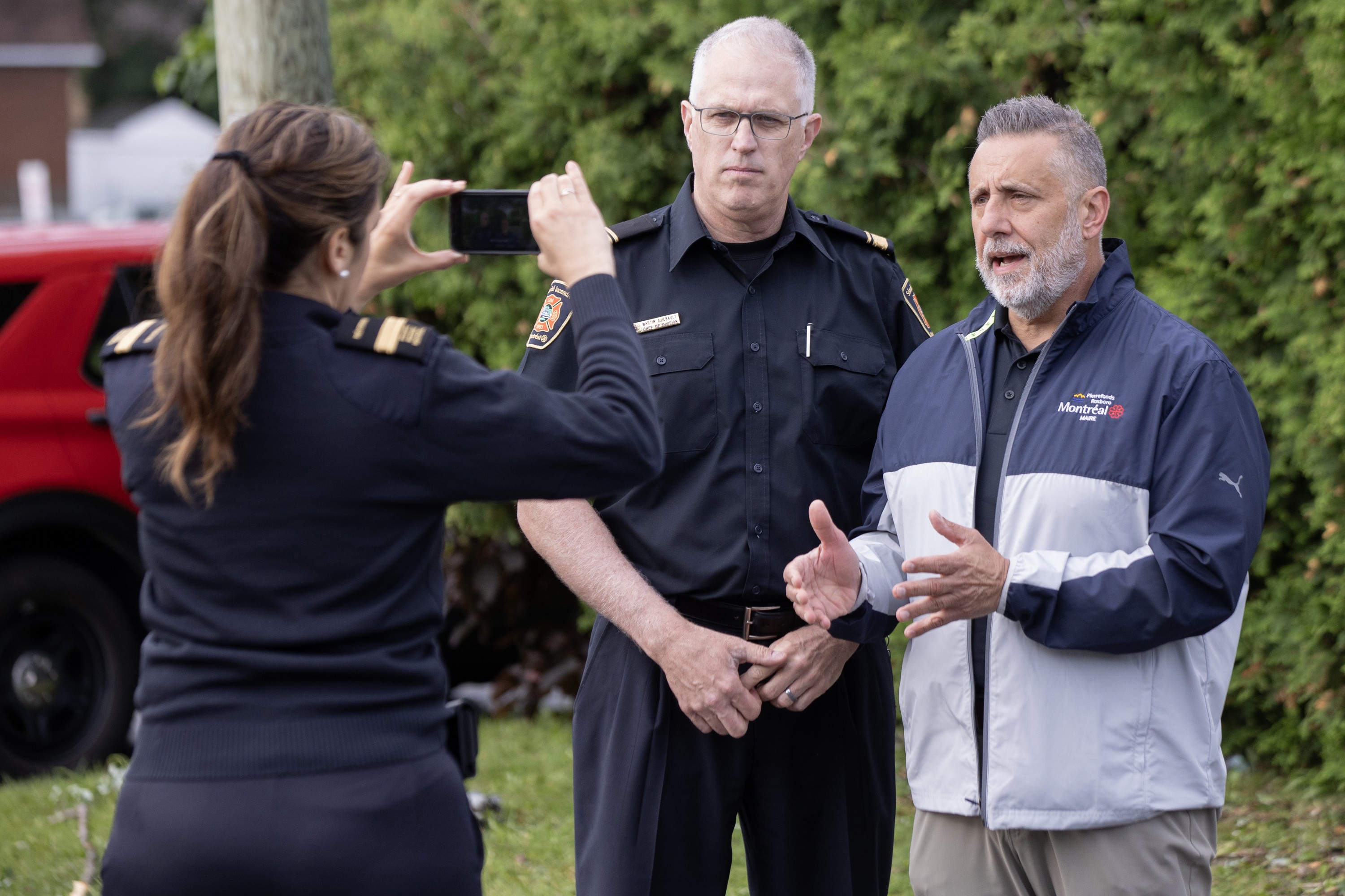 Pierrefonds-Roxboro borough mayor Jim Beis speaks next to a Montreal fire department spokesperson as an official films them on her phone.