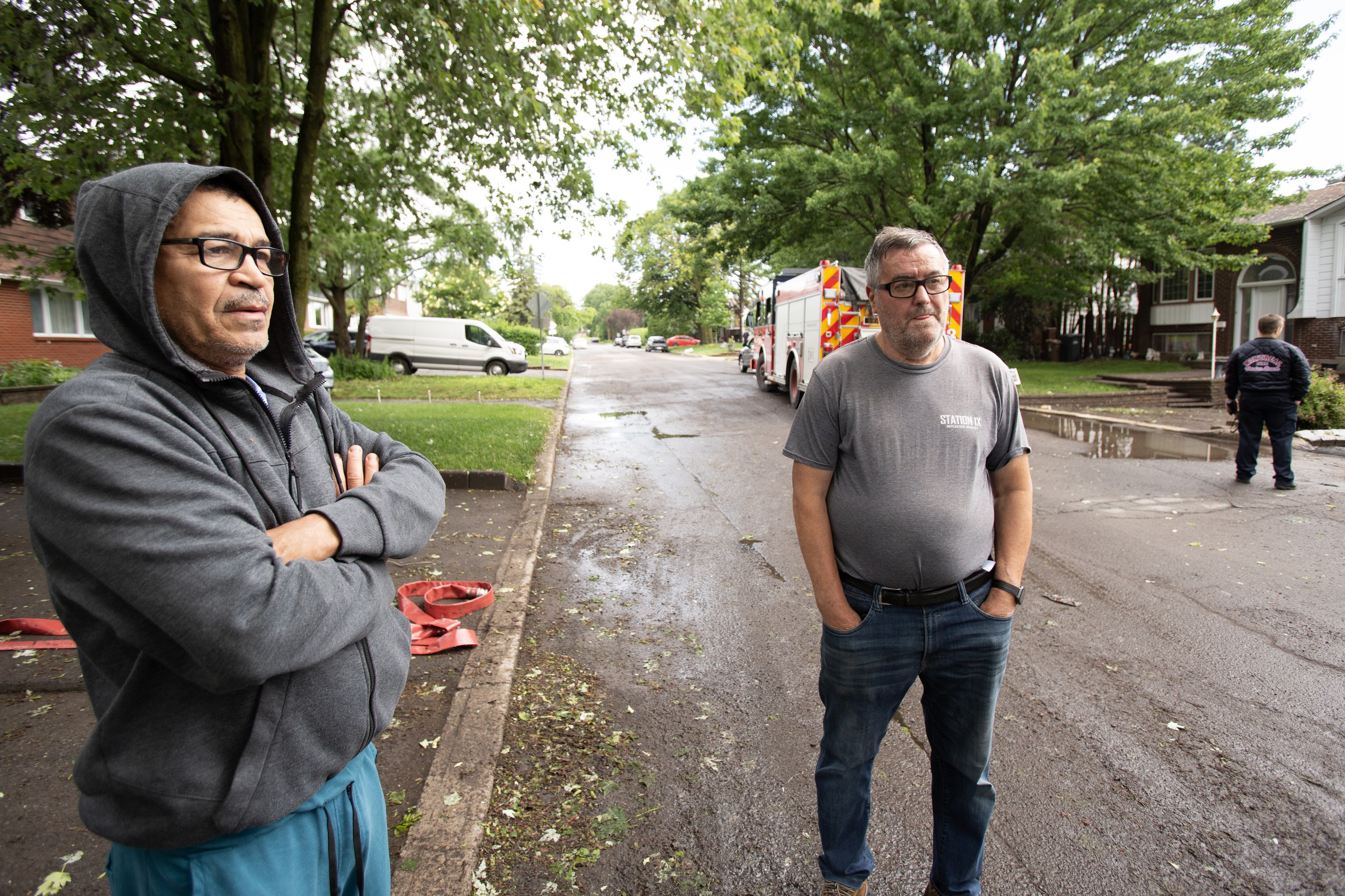 Two men stand on a street with a fire truck in the background.