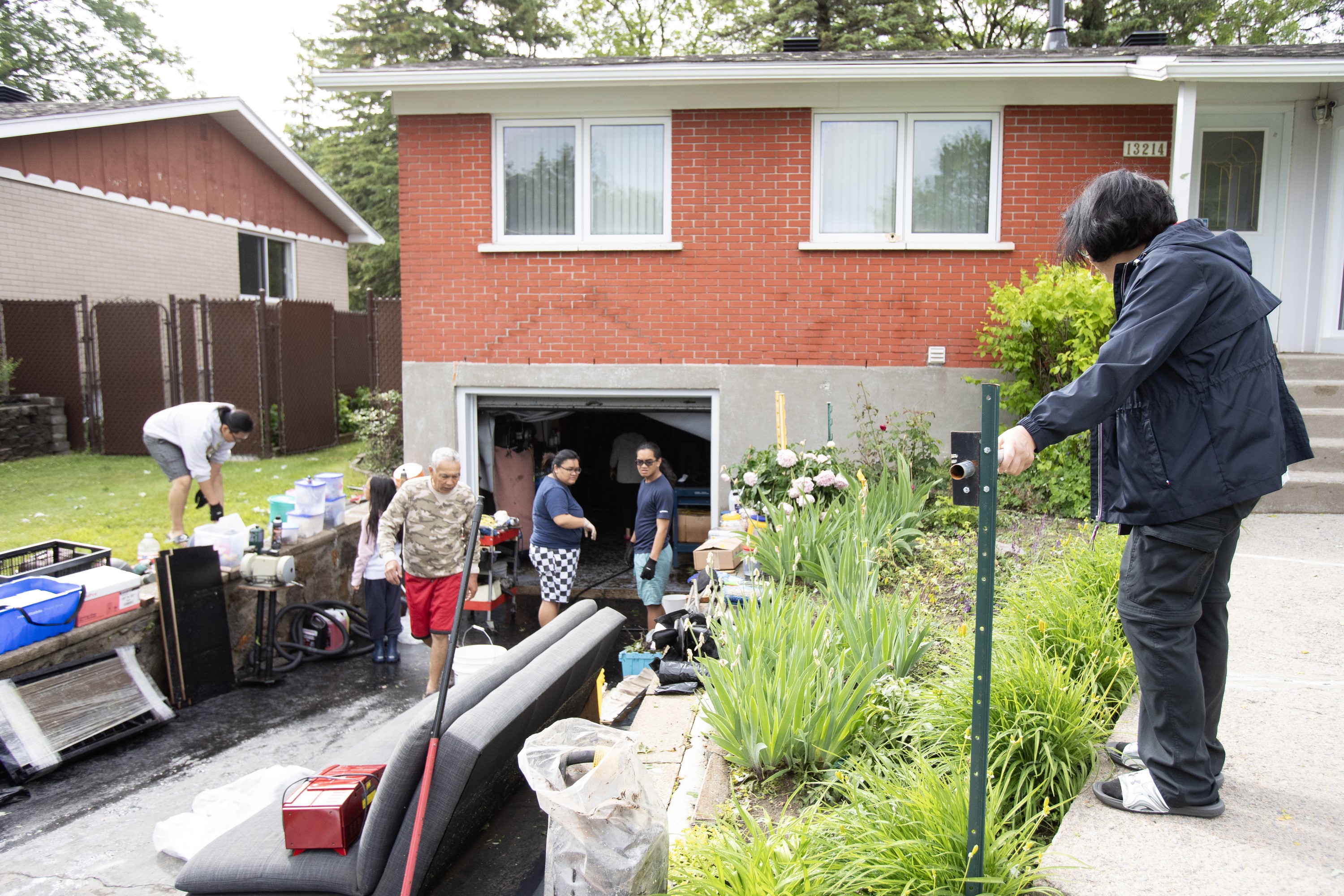 A woman watches from a walkway outside a house as other people sort through belongings in a driveway.