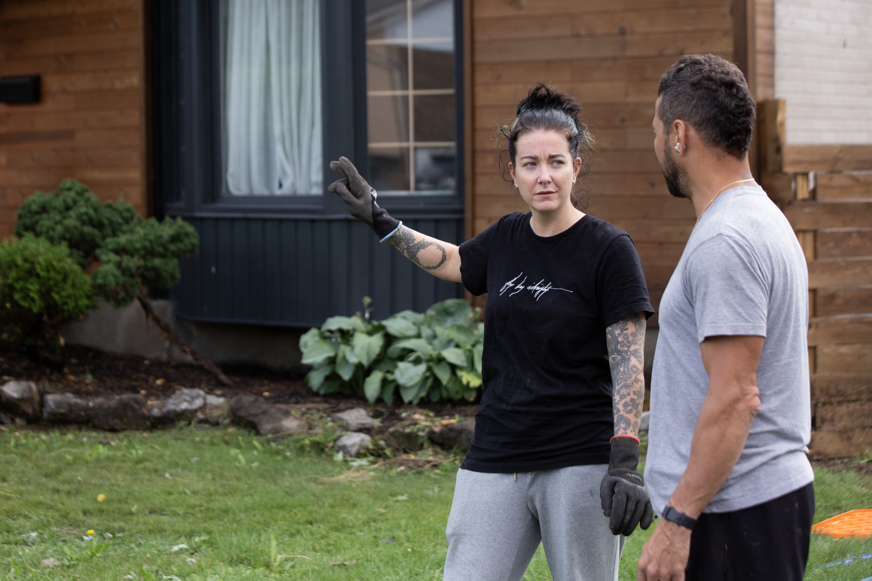 A woman wearing gloves gestures while she speaks to a man outside a house.
