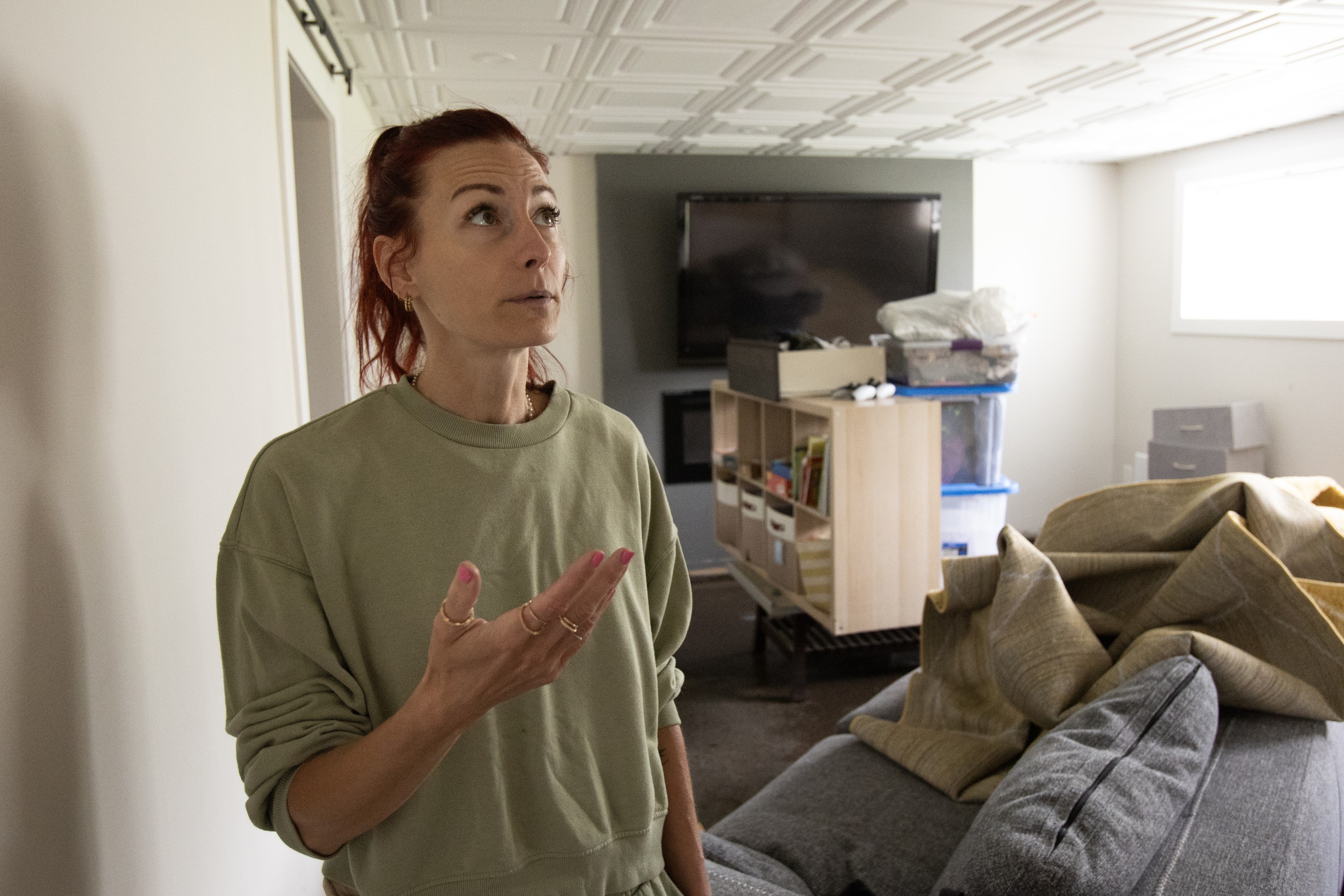 A woman gestures and looks up in a basement room, with possessions stacked behind her in the wake of flooding.