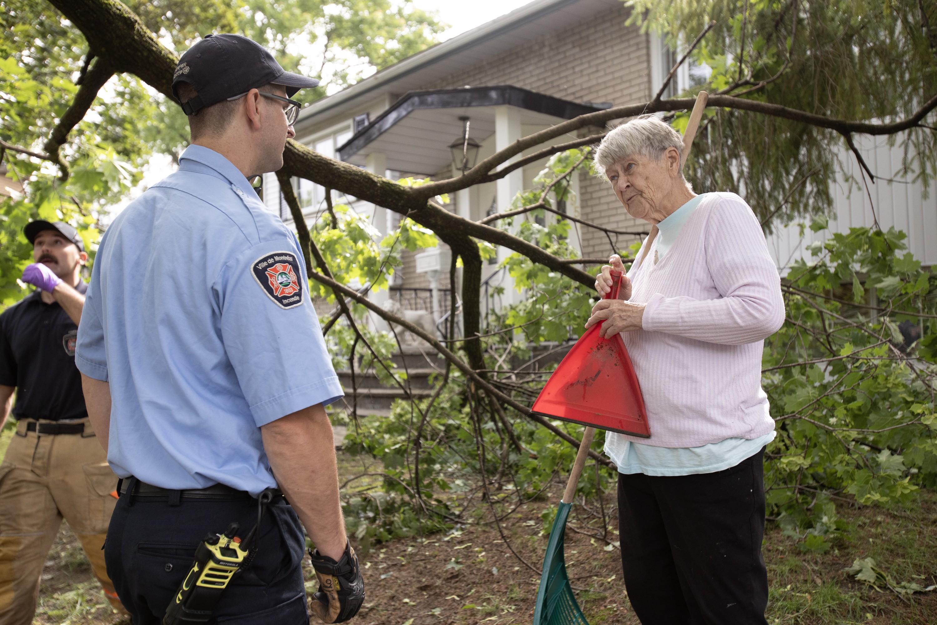 A woman holds a dustpan and rake as she speaks to a firefighter on a lawn in front of a damaged tree branch.