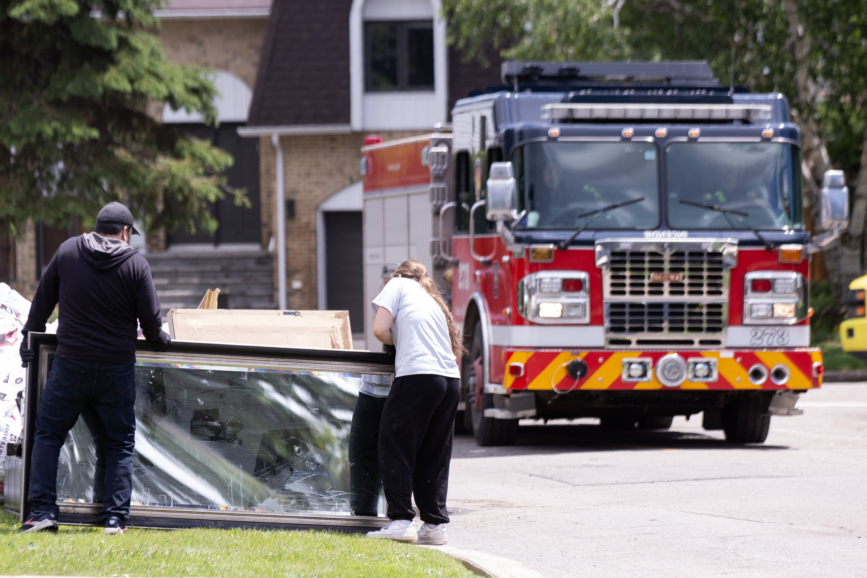 Two people are seen from behind as they bring flood-damaged household items to the curb. A fire truck is in the background.