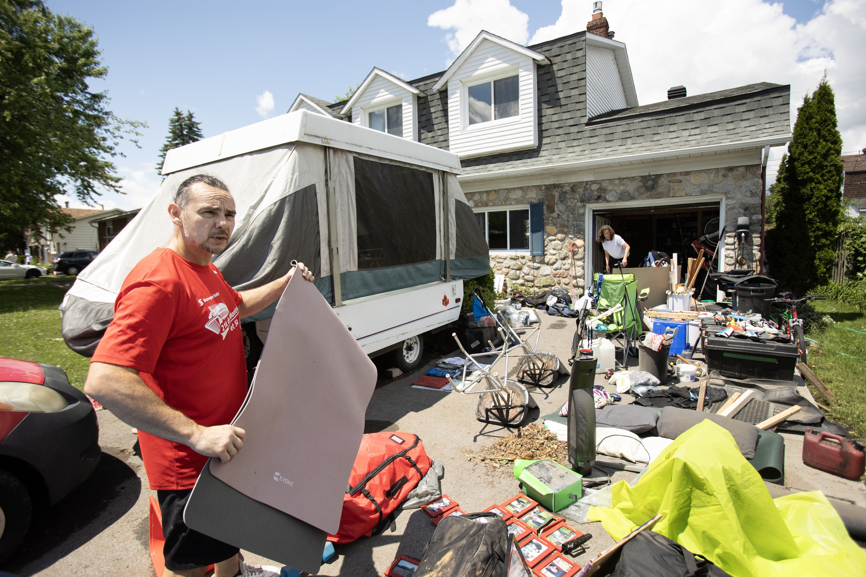 A man carries household items on a driveway strewn with flood-damaged possessions in front of a house. A woman works in the garage of the house in the background.