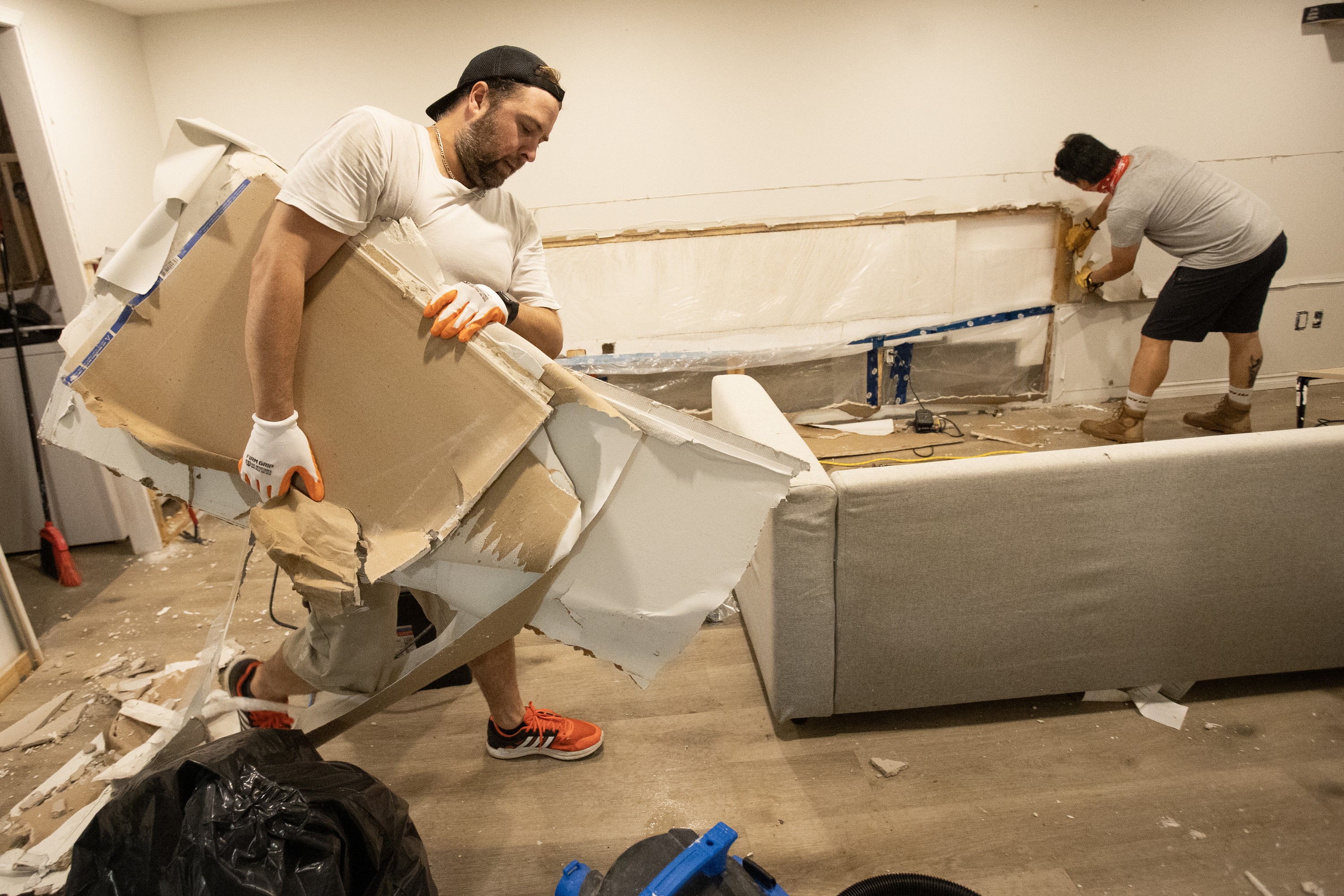A man carries a stack of damaged drywall as another man pulls pieces from the wall in the background.