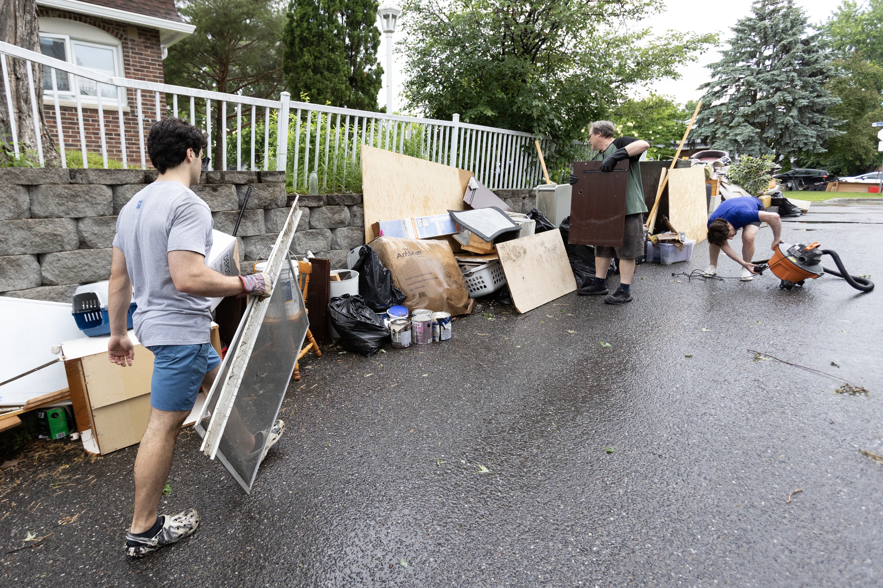 Two people bring flood-damaged belongings to the side of a road outside a house.