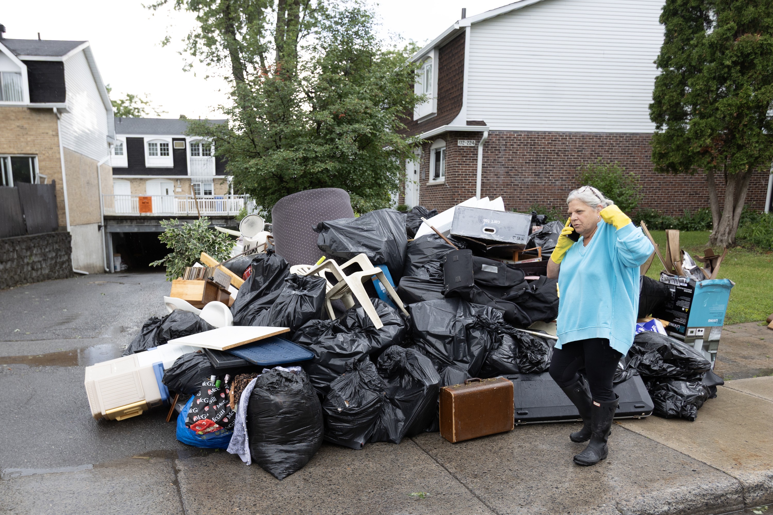 A woman wearing yellow cleaning gloves walks past a large pile of debris and damaged belongings at the end of a driveway.