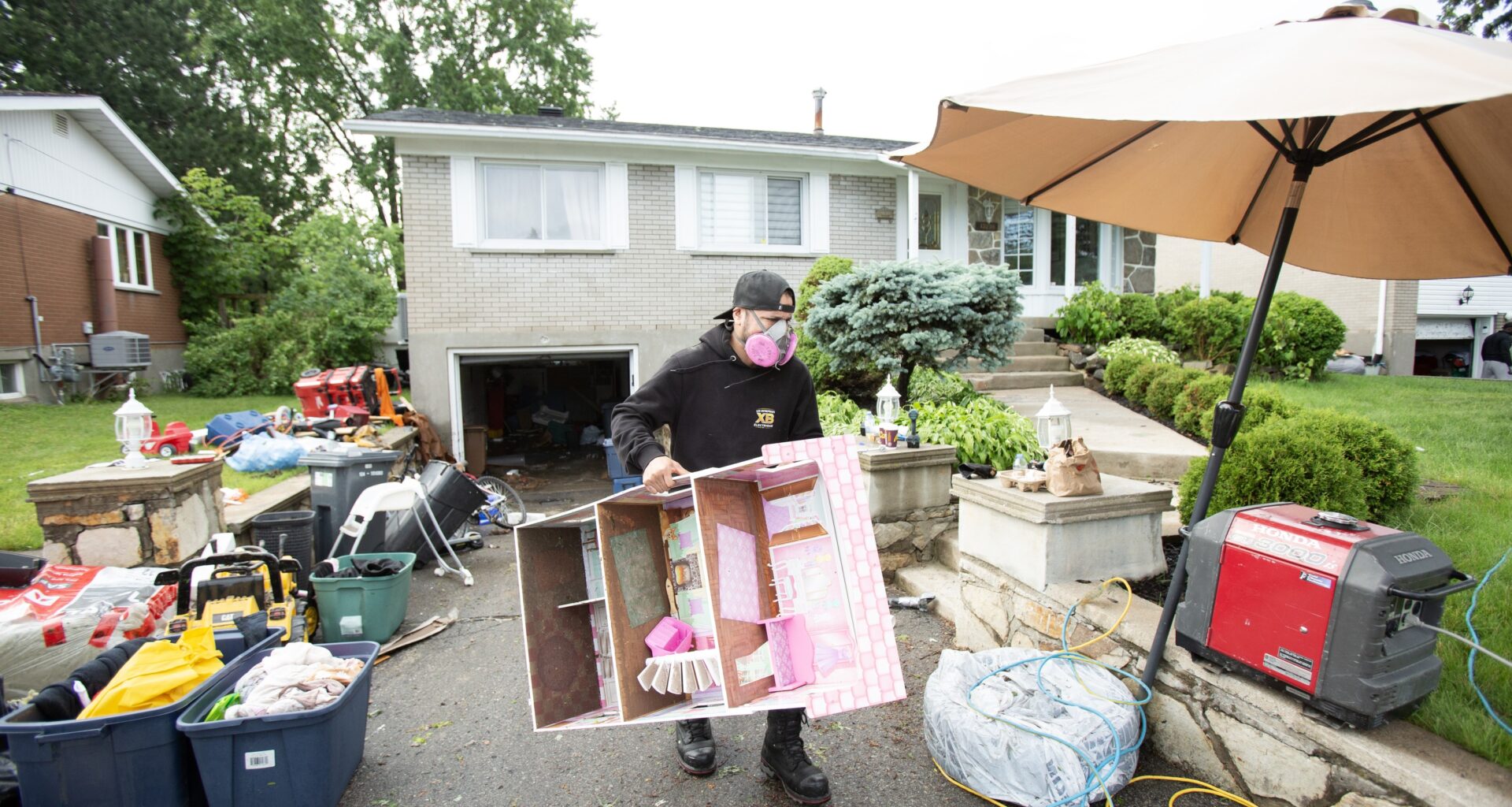 A man wearing a mask carries a doll house up a driveway strewn with belongings after flooding.
