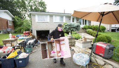 A man wearing a mask carries a doll house up a driveway strewn with belongings after flooding.