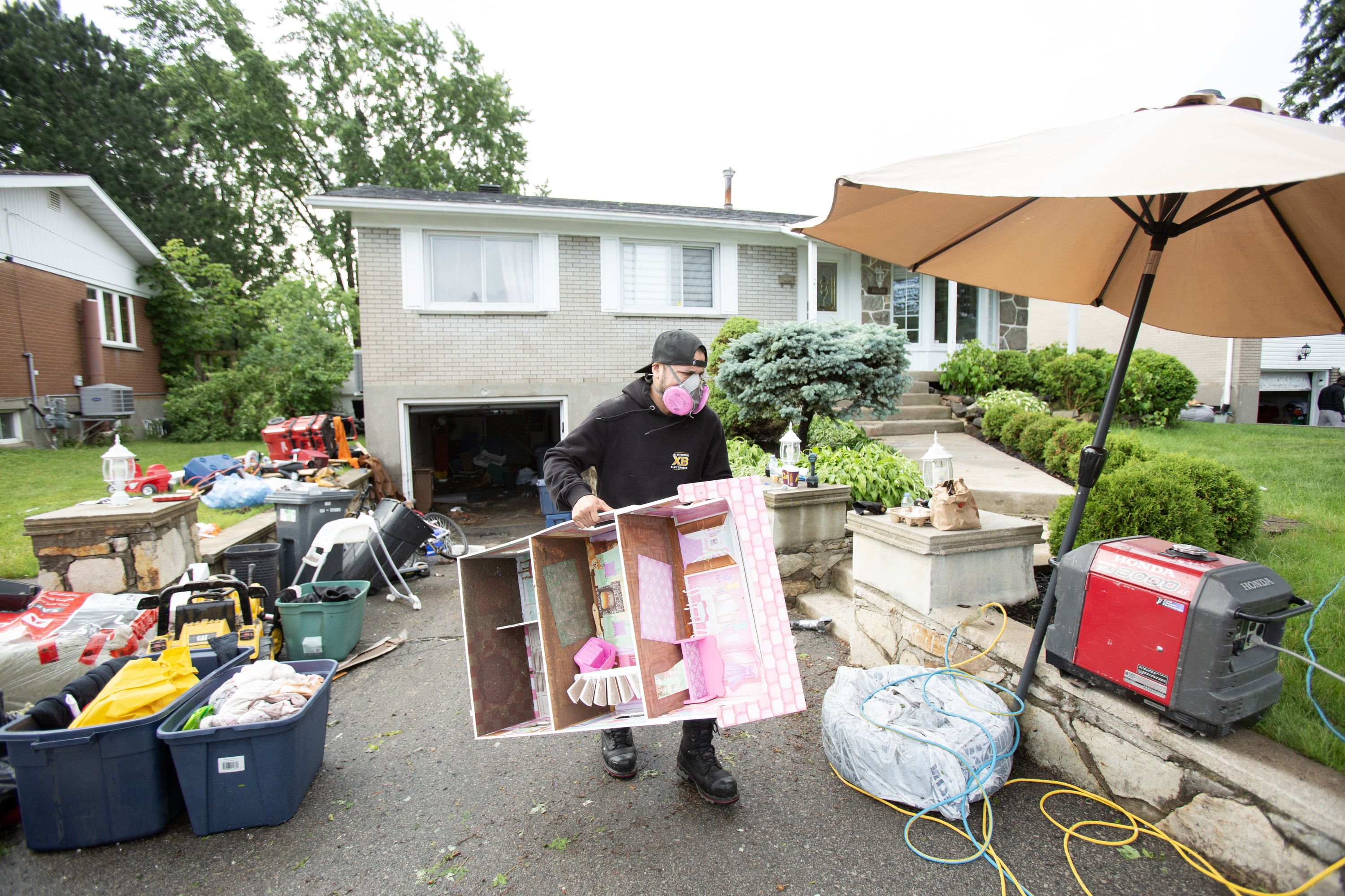 A man wearing a mask carries a doll house up a driveway strewn with belongings after flooding.