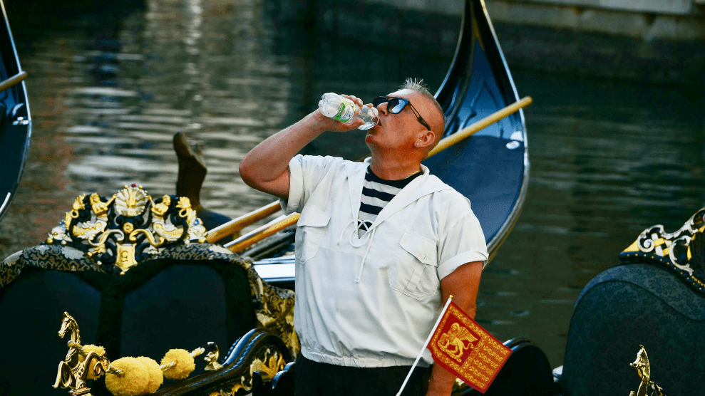 A gondolier man in white shirt drinks from plastic water bottle on top of black and gold gondolas.
