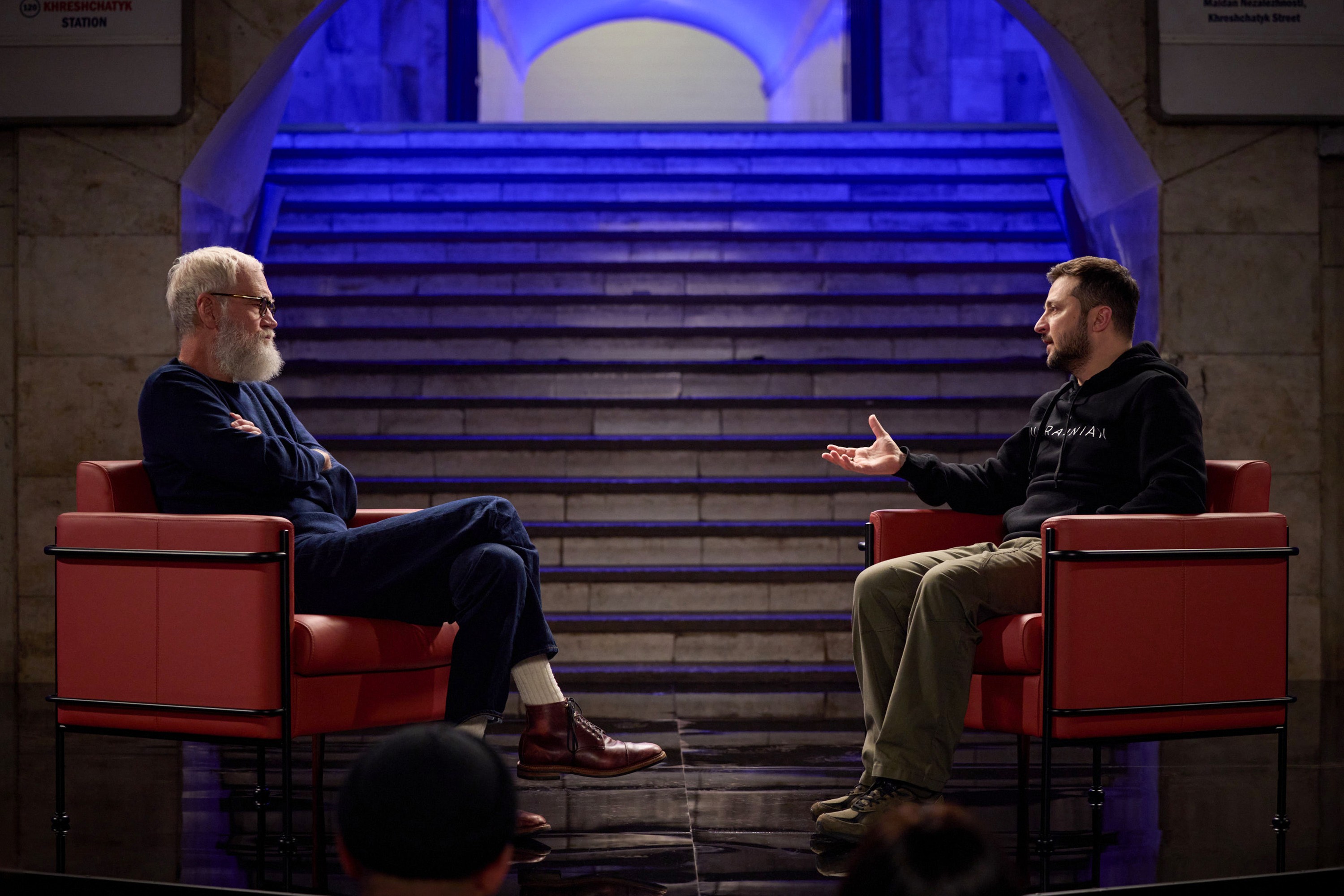 David Letterman sits in a chair with arms crossed, listening to Ukrainian President Volodymyr Zelenskyy, who sits opposite him while gesturing.