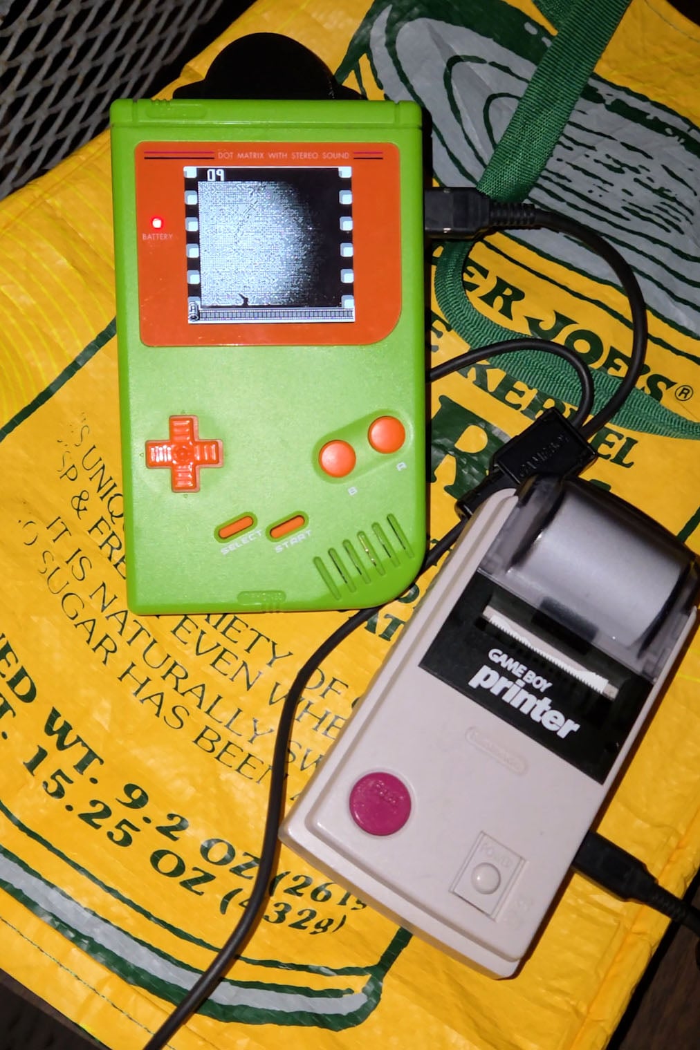A green and orange handheld gaming device is connected to a gray Game Boy Printer, both resting on a yellow Trader Joe’s shopping bag. The devices are lit by a camera flash.