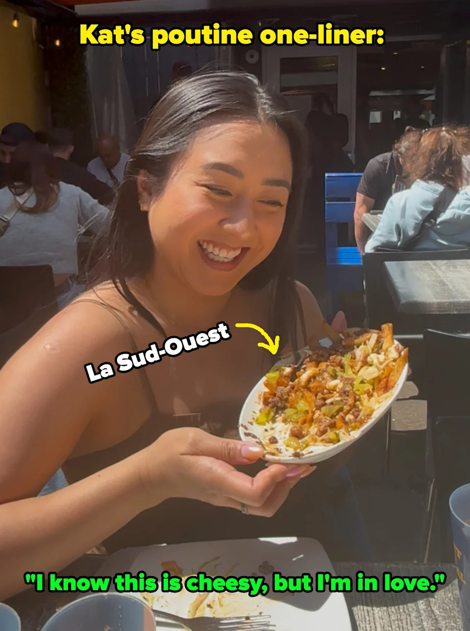 Person with short hair sitting outdoors, holding a plate of food, smiling gladly at it. People are seated at tables in the background