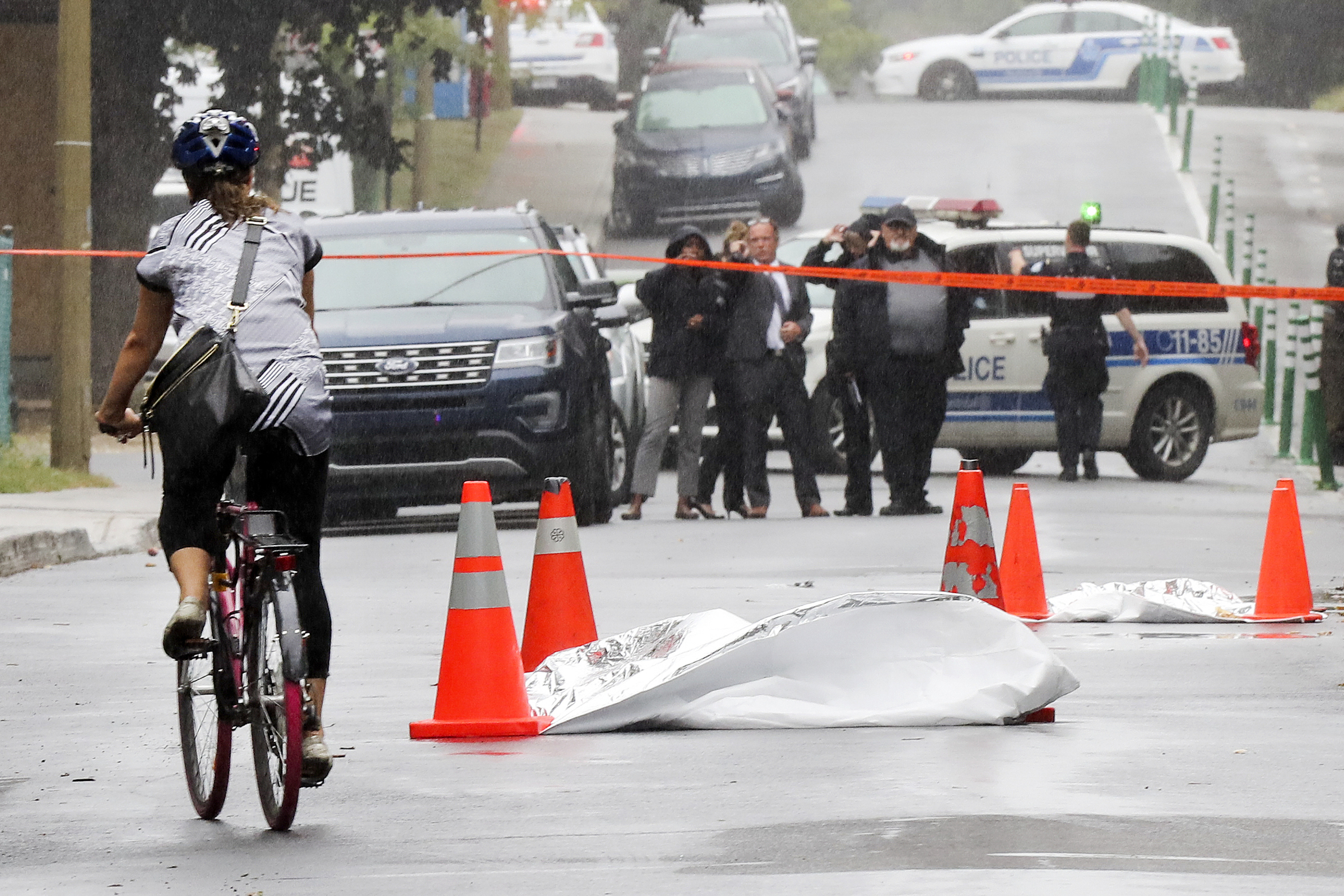 A body bag lies in the street with orange cones around it as a cyclist goes by, with police officers in the distant background.
