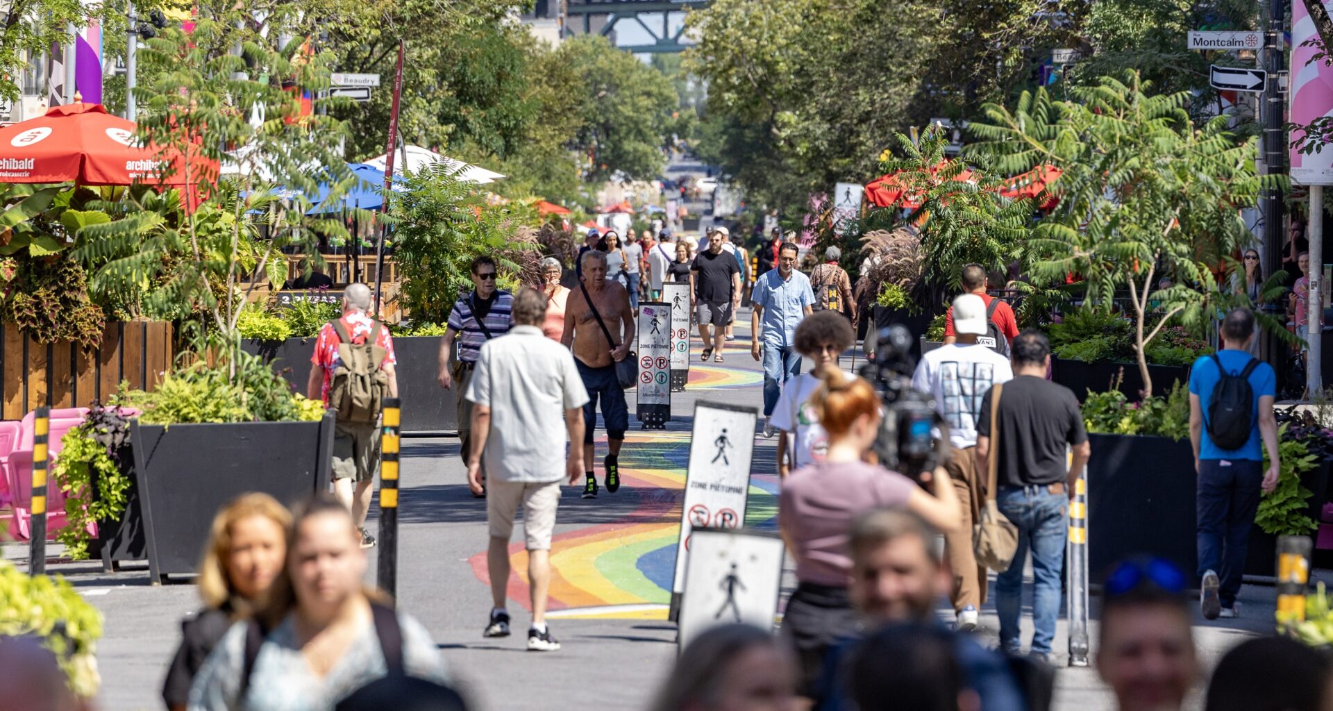 Dozens of people crowd onto a pedestrian-only street lined with restaurants and trees. There's a rainbow path painted on the street.