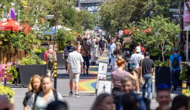 Dozens of people crowd onto a pedestrian-only street lined with restaurants and trees. There's a rainbow path painted on the street.