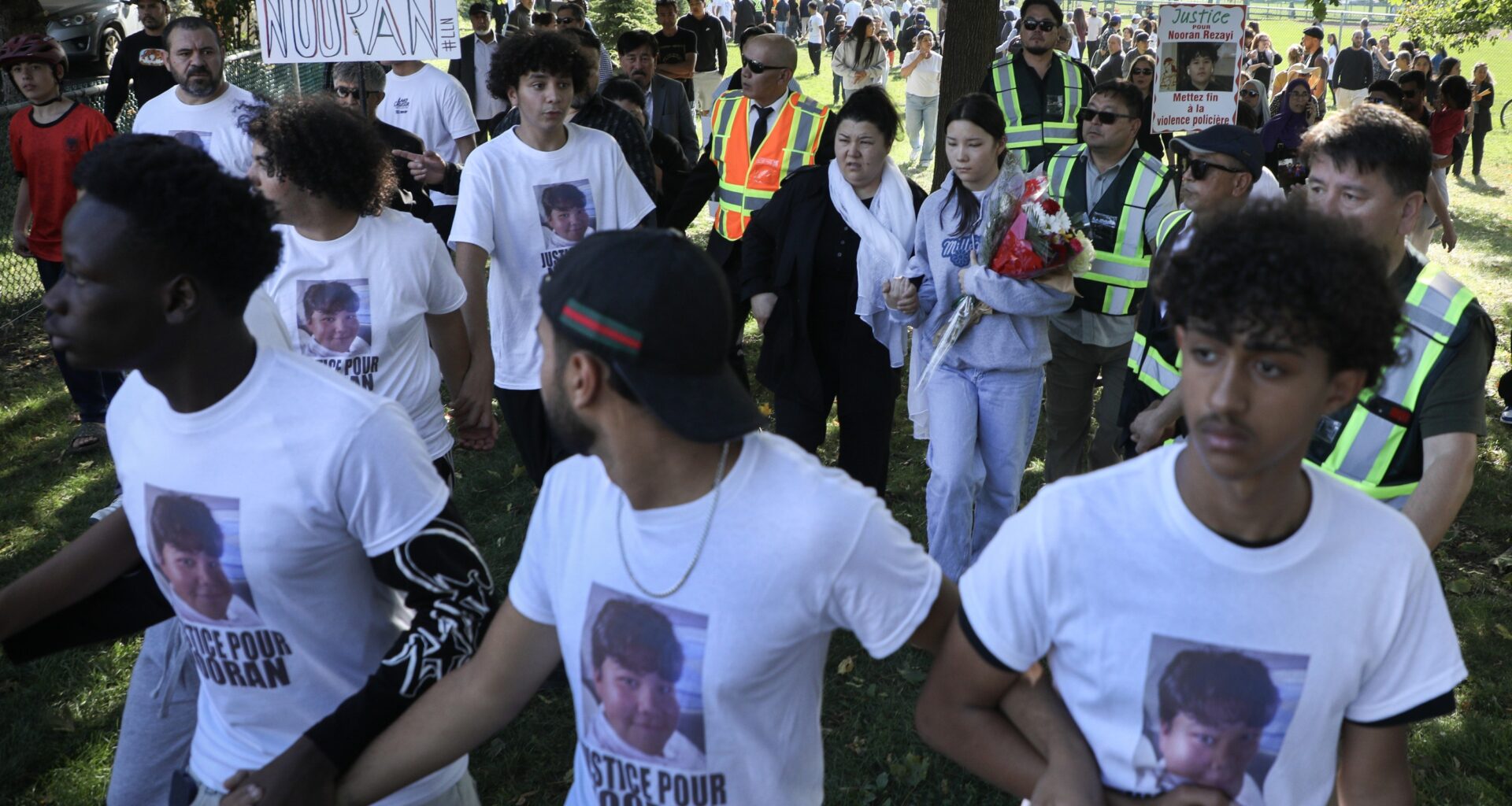 Three young men link arms and walk with a crowd of demonstrators behind them. A placard reads Justice pour notre frere Nooran