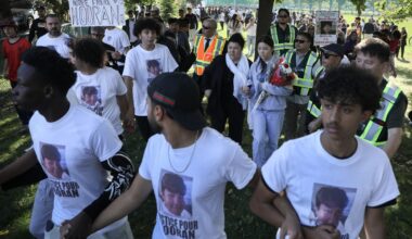 Three young men link arms and walk with a crowd of demonstrators behind them. A placard reads Justice pour notre frere Nooran