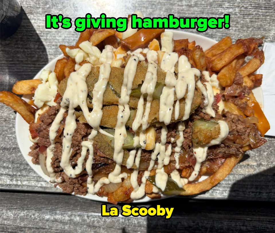 A plate of loaded fries topped with cheese curds, roast beef, fried pickles, and drizzled with a creamy sauce on a wooden table