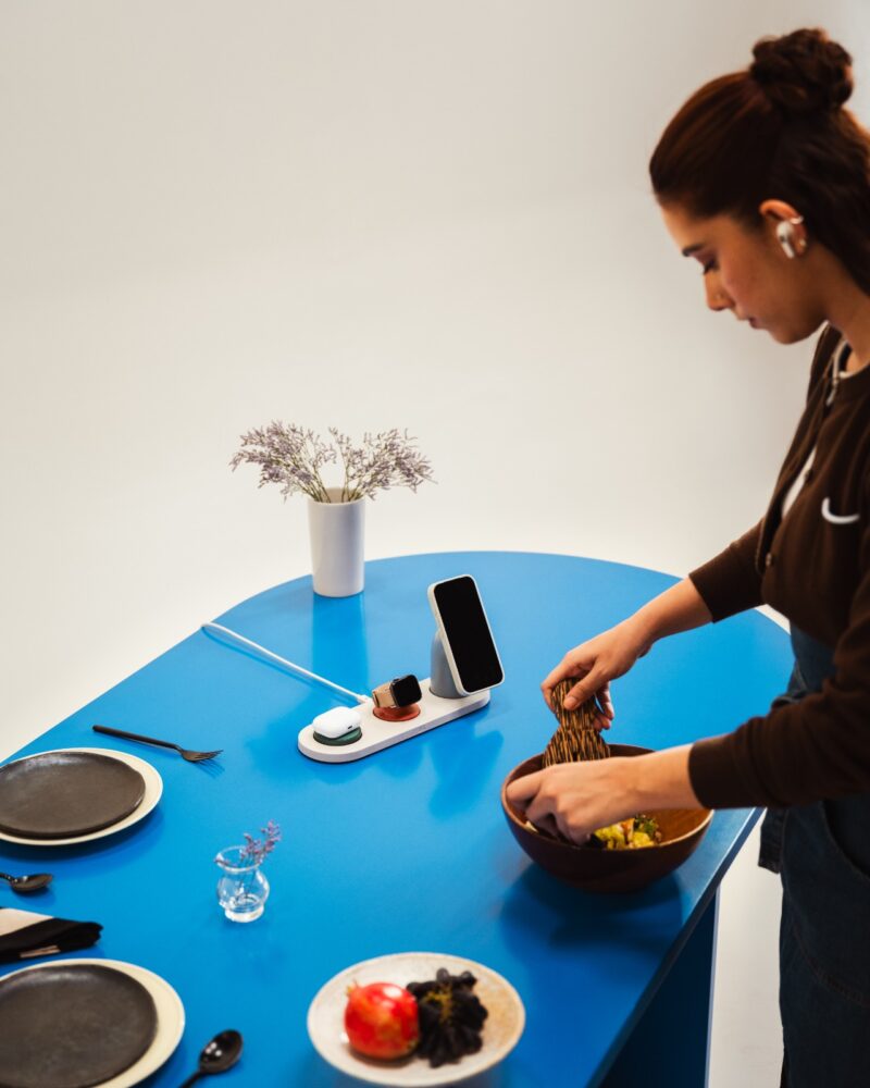 A woman prepares food at a blue table set with plates, utensils, a small vase with flowers, and a DailyObjects smartphone stand.