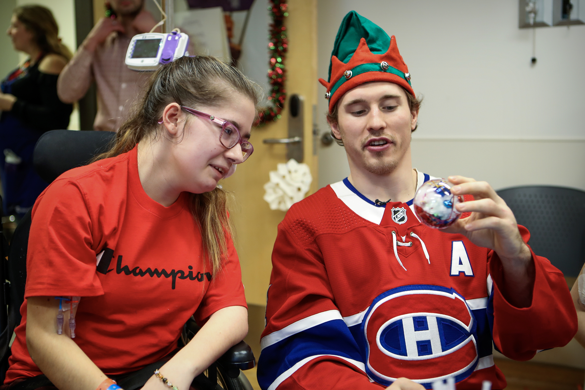 Brendan Gallagher holds up a Christmas ornament while wearing an elf hat and Canadiens jersey sitting next to a young woman in a hospital.